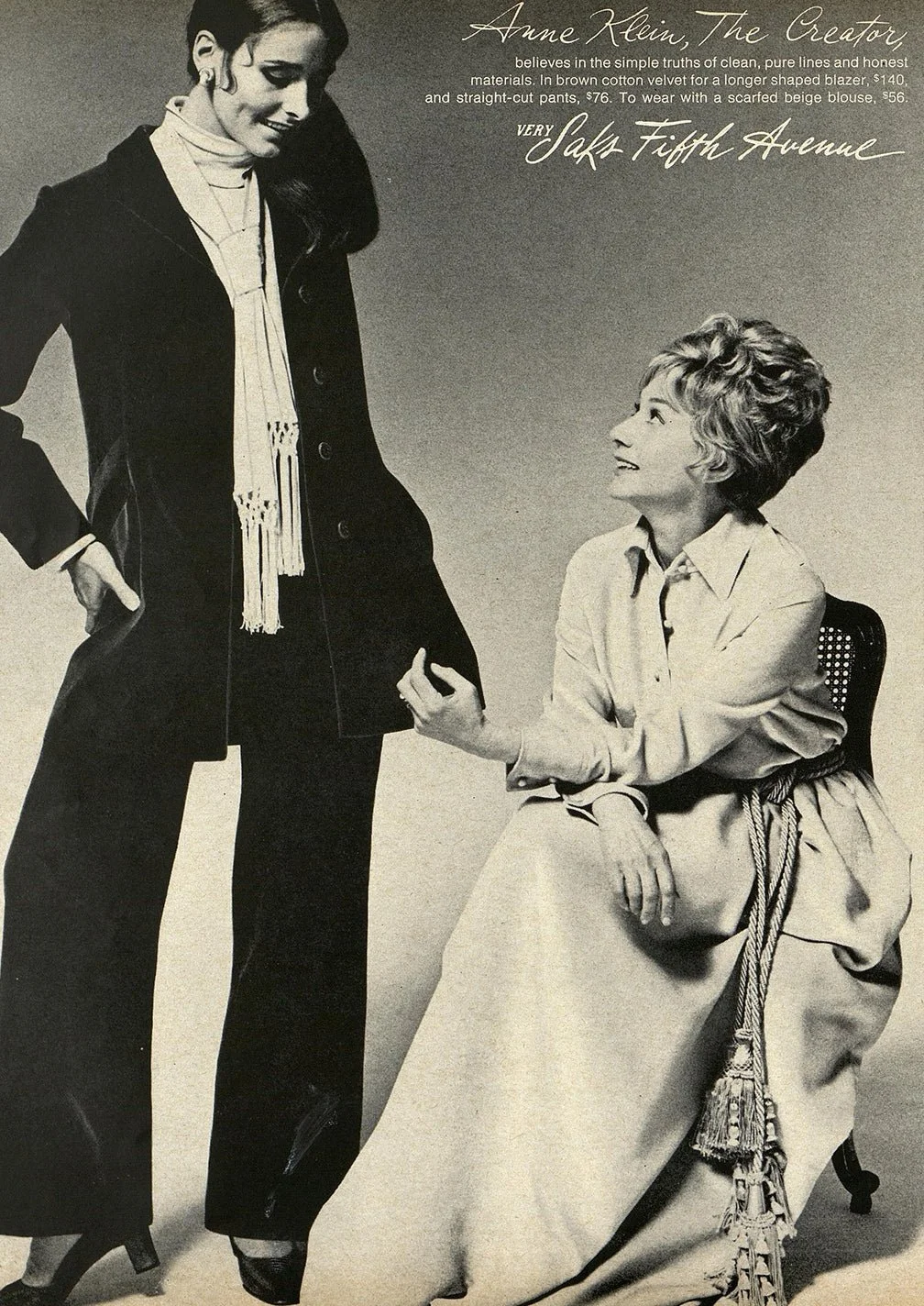 Black and white photograph of two women, one standing and the other seated. The standing woman is smiling, wearing a dark blazer, pants, and a scarf, with her hand on her hip. The seated woman is looking up at her, wearing a light-colored blouse and a long skirt with a decorative rope belt, and is holding the standing woman's hand.