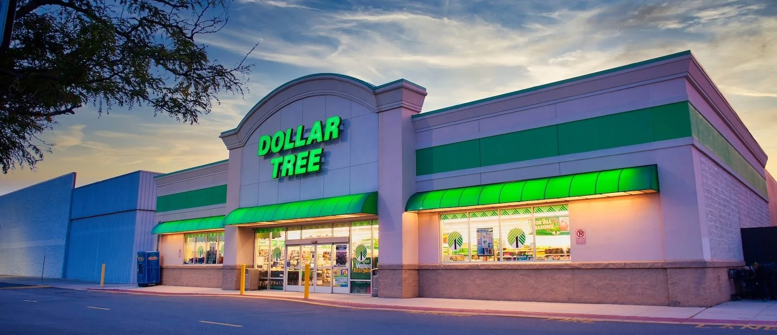 Exterior of a Dollar Tree store at dusk with a green sign and green awnings, large windows, and a parking lot in front.