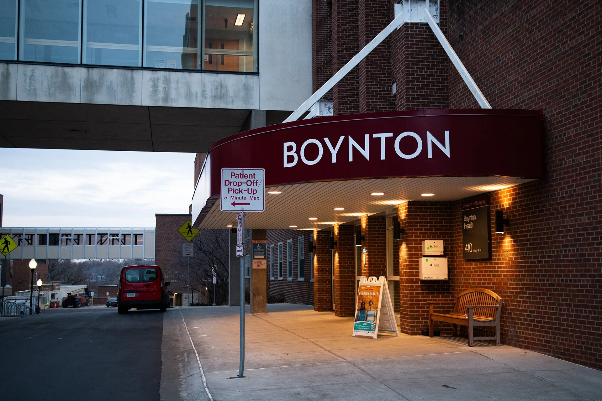Building entrance with a maroon awning labeled 'BOYNTON', a bench, a sign for patient drop-off and pick-up, and a sidewalk leading to a street with parked cars and streetlights at dusk.