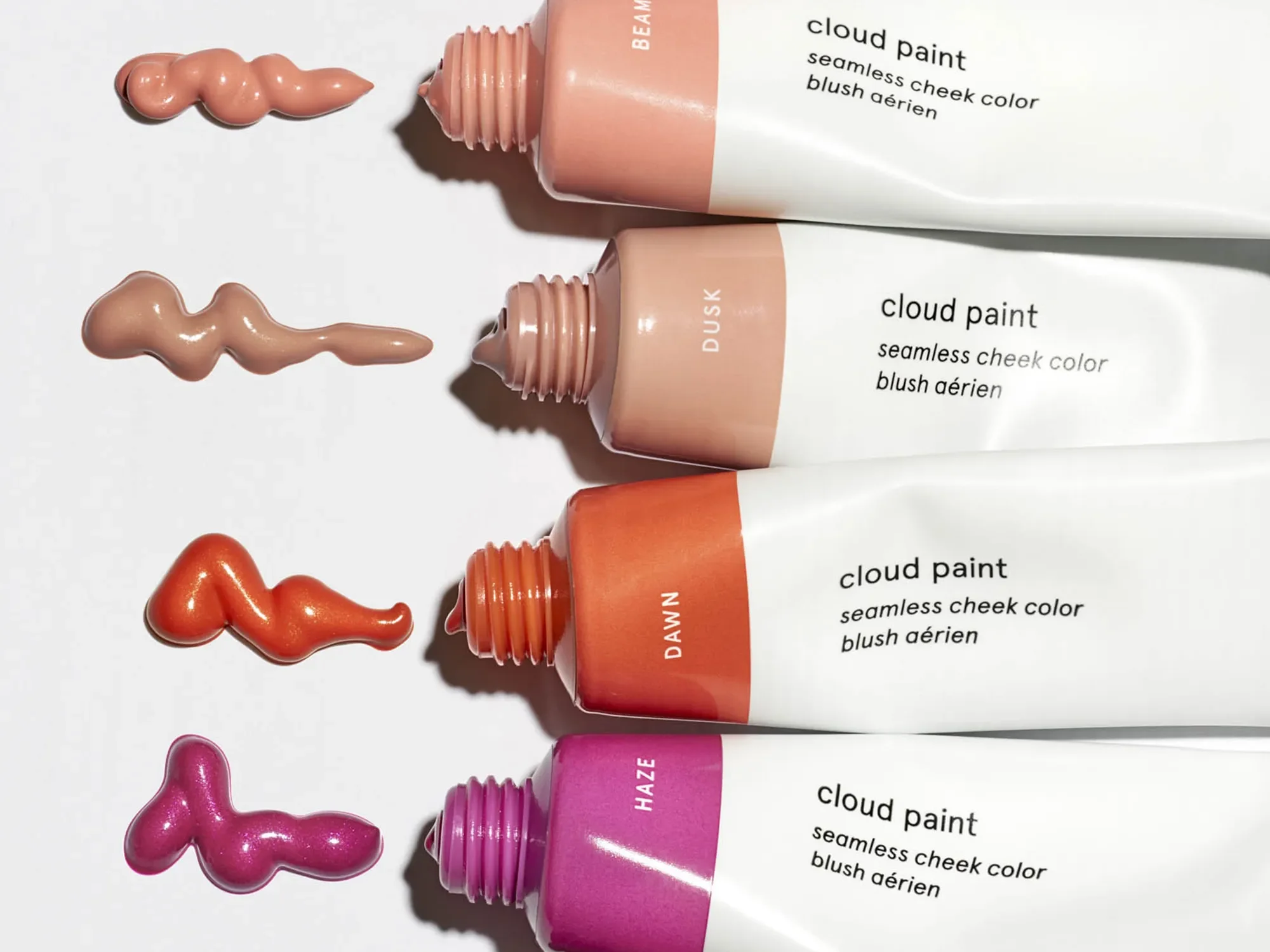 Four tubes of cloud paint cheek color from Glossier in different shades, with each tube squeezed out to show the color: top tube labeled 'BEAM' with light pink color, second tube labeled 'DUSK' with beige pink color, third tube labeled 'DAWN' with coral color, bottom tube labeled 'HAZE' with vibrant pink color.