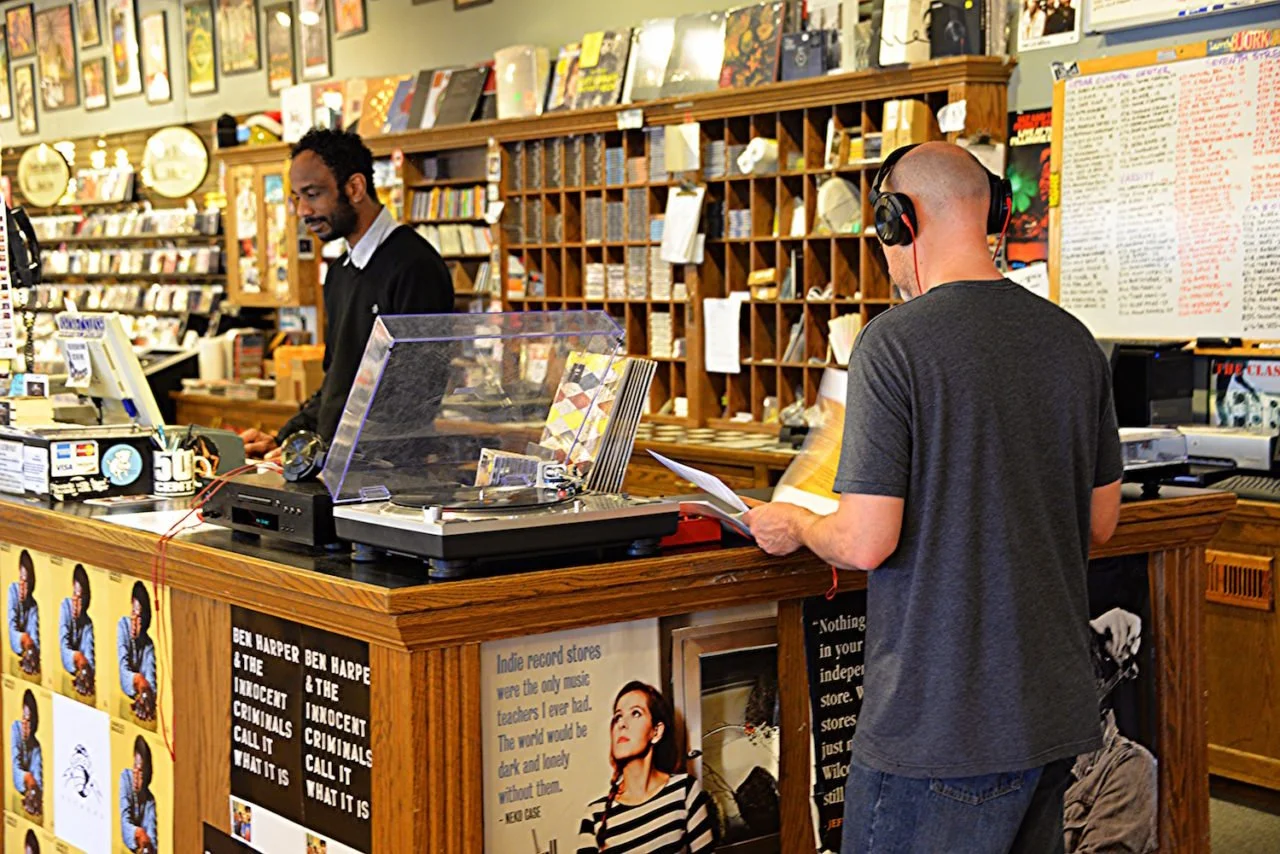 Two men in a record store. One man is working behind the counter, and the other is listening to music with headphones on while holding a paper. The store has shelves filled with records and posters on the walls.