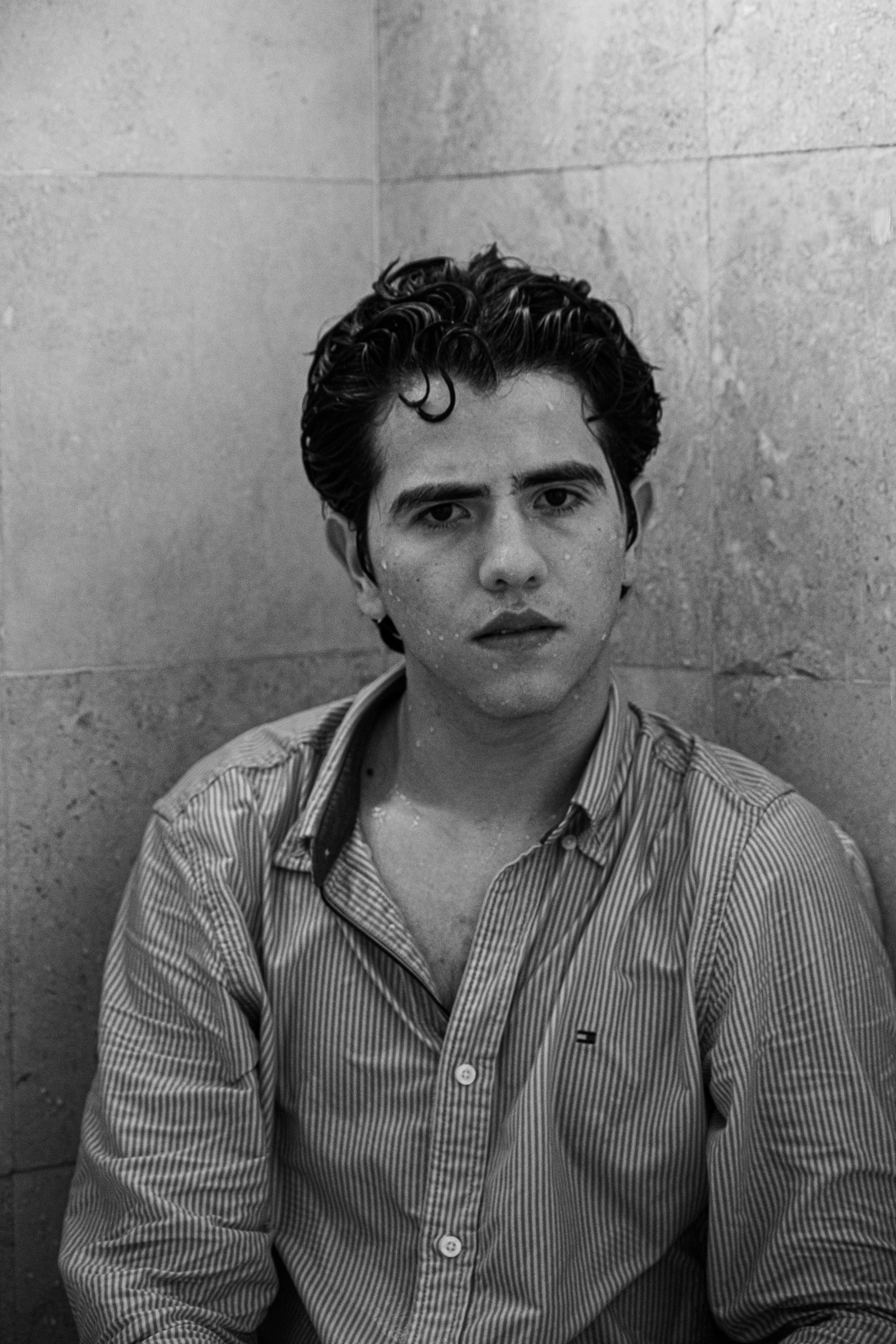 Black and white photo of Ray Maldonado with wet hair and water droplets on face, wearing a striped button-up shirt, sitting against a tiled wall, looking directly at the camera.