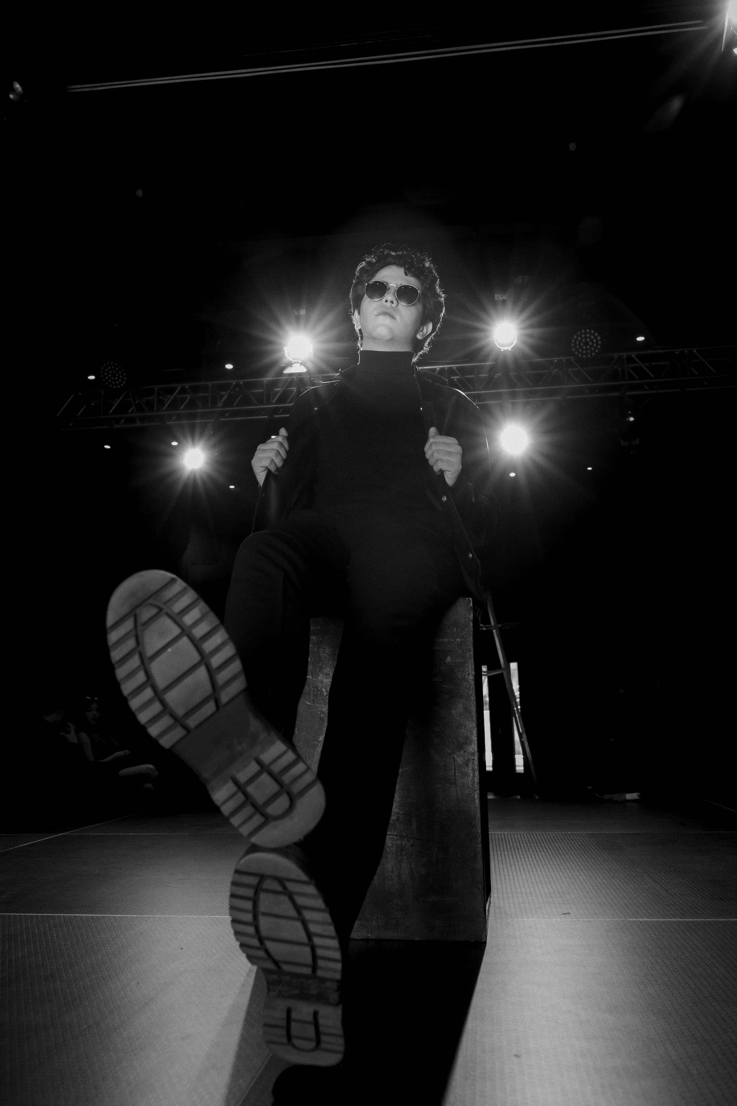 Ray Maldonado with sunglasses sits on a platform with one foot raised, looking confident, with stage lights shining behind them in a dark environment.