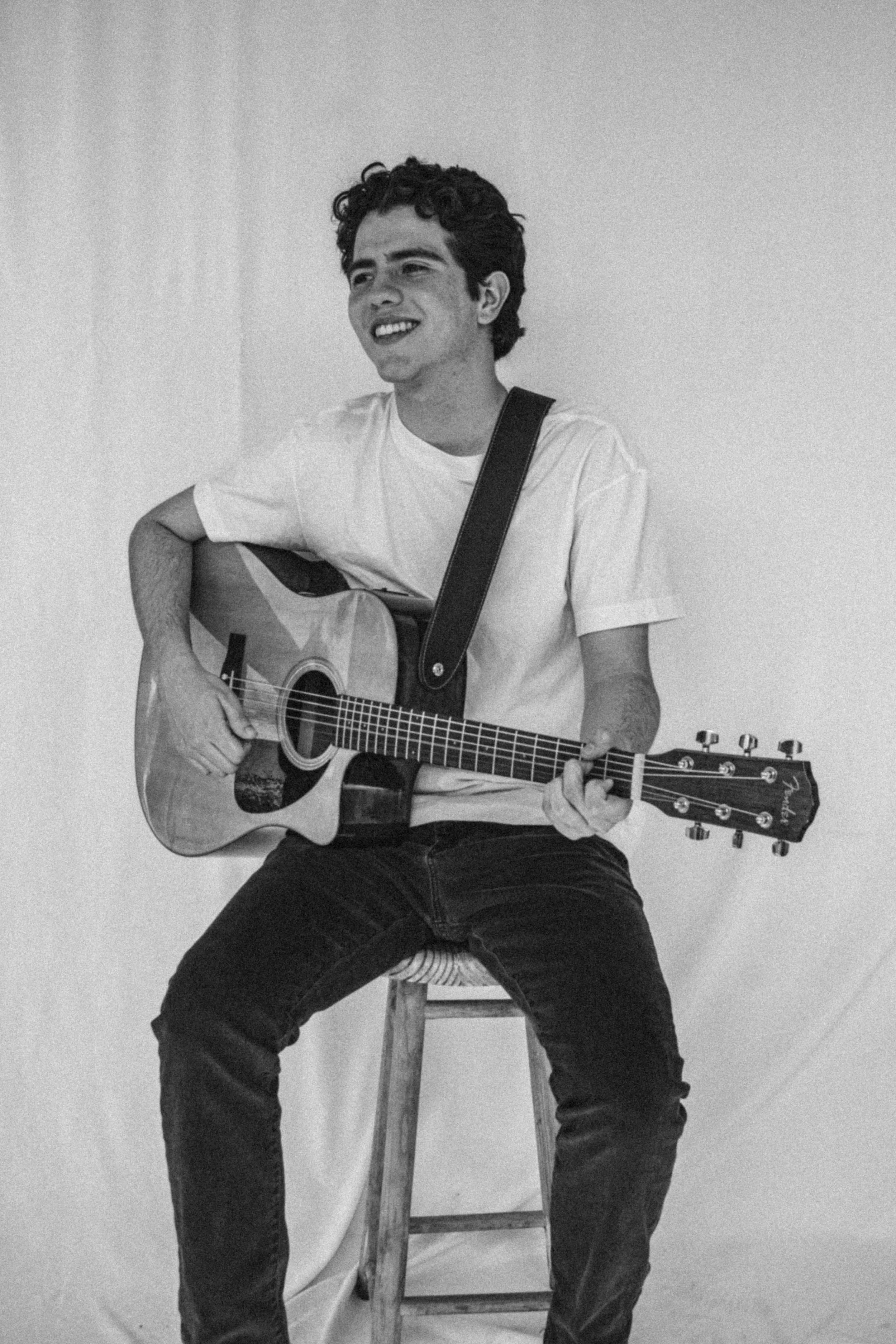 Ray Maldonado playing an acoustic guitar while sitting on a stool, smiling, against a plain background.