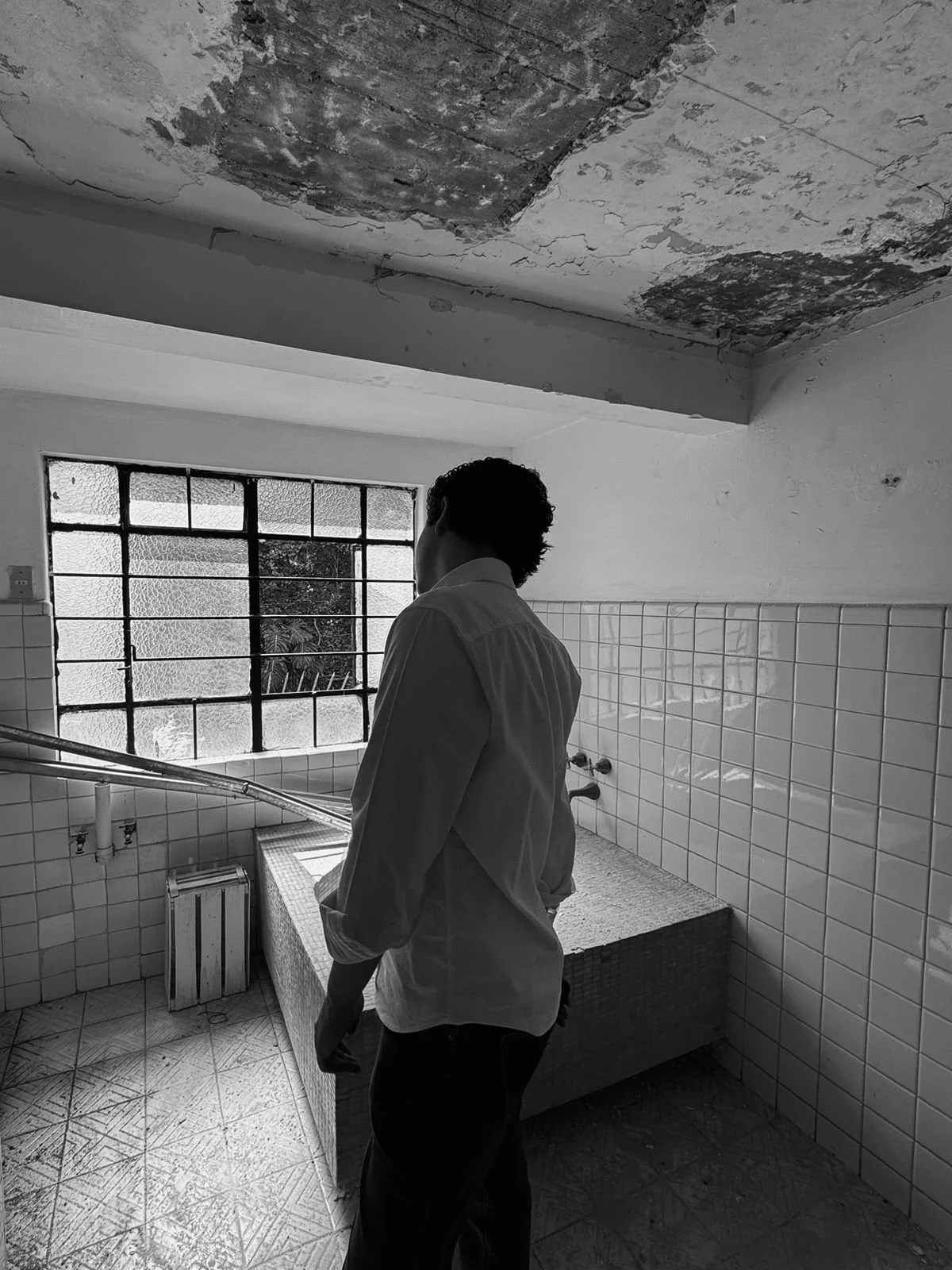 Ray Maldonado standing in a dilapidated bathroom with peeling paint on the ceiling, looking away from the camera, with a large window and tiled walls.