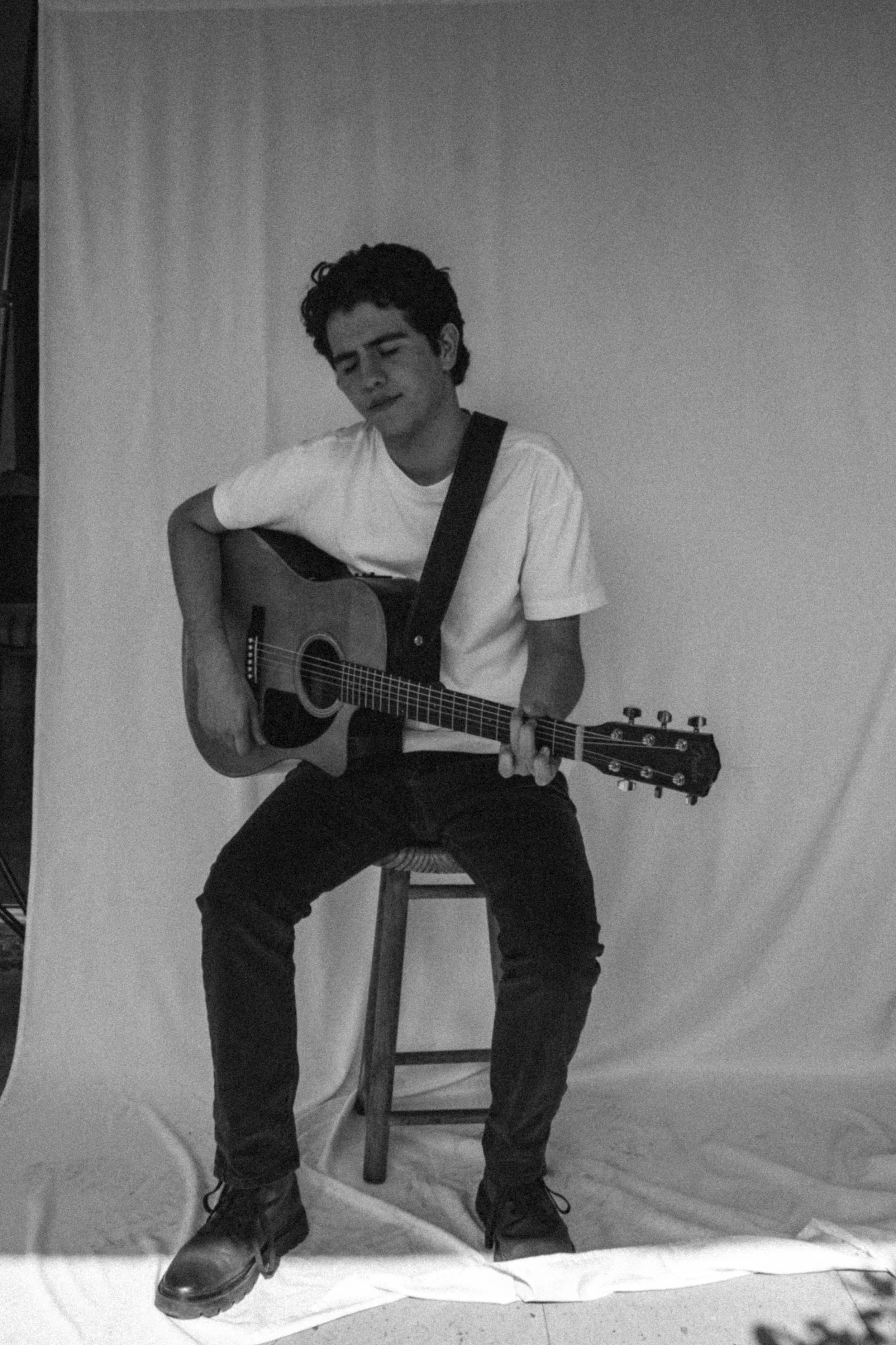 A young man is sitting on a wooden stool, playing an acoustic guitar. He is wearing a white t-shirt, black pants, and black shoes. His eyes are closed, and he appears to be deeply immersed in the music. The background is a plain, light-colored curtain.