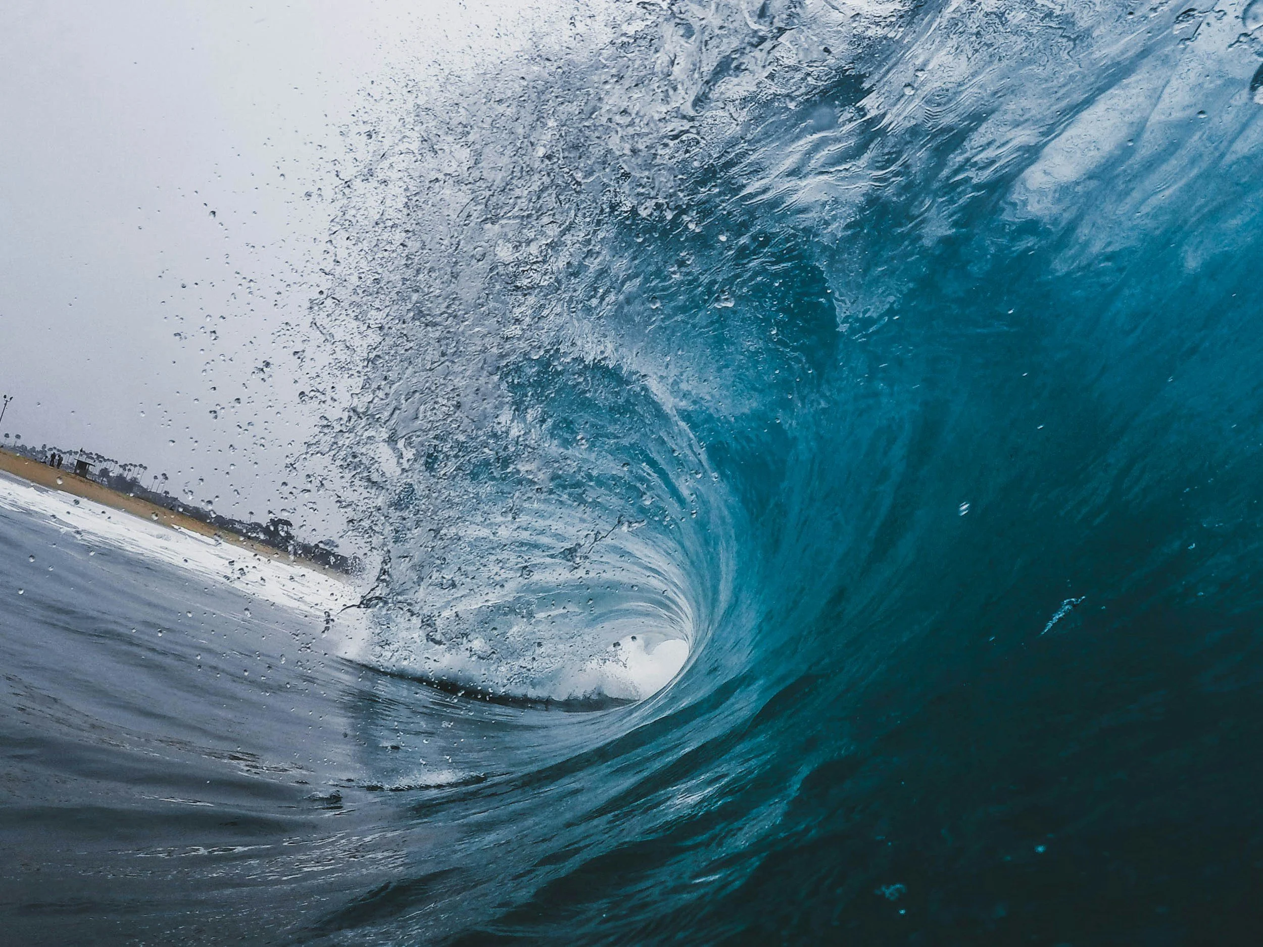 A large ocean wave curling over with water splashing, viewed from inside or near the wave, with a distant shoreline and cloudy sky in the background.