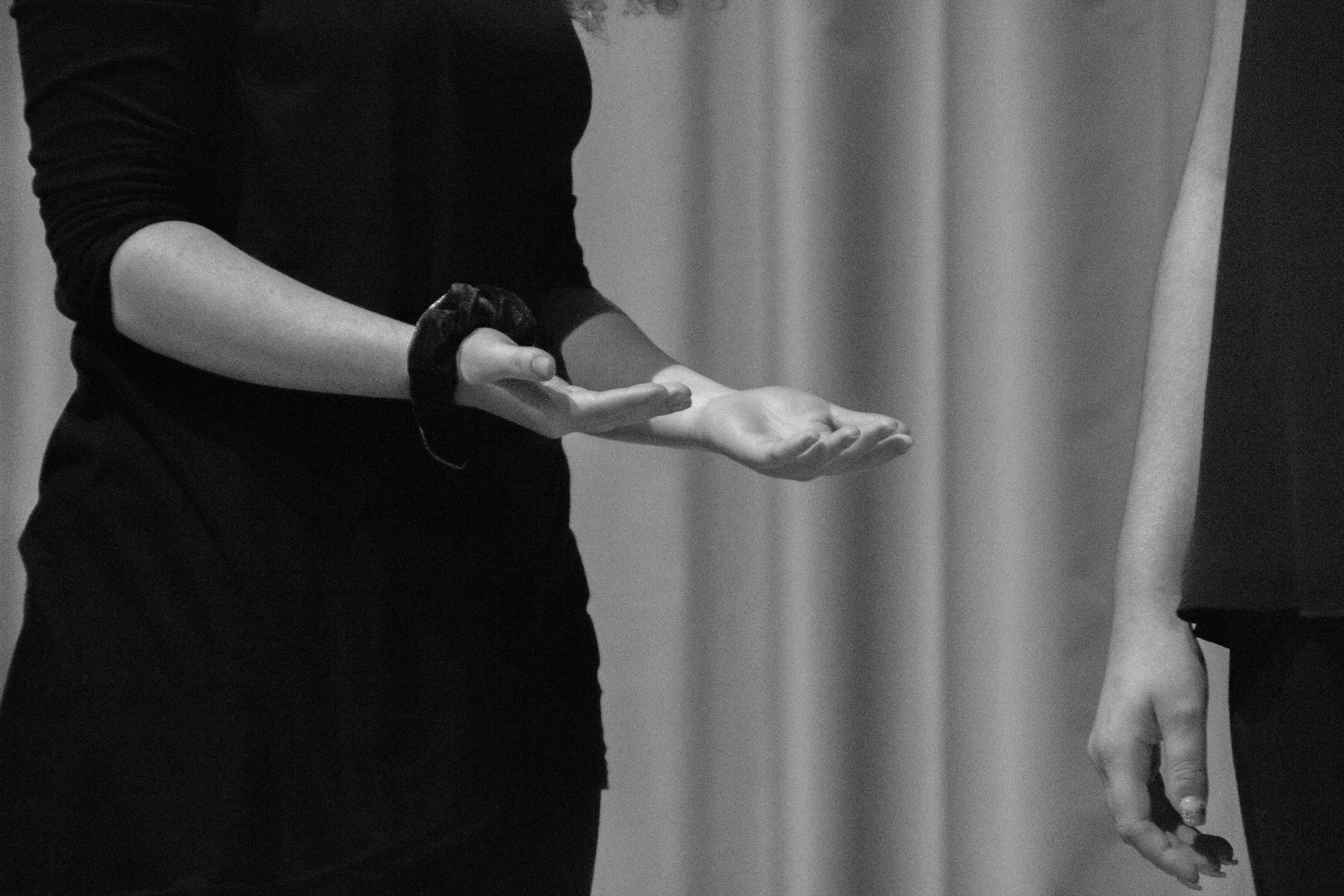 A black and white photo of a woman gesturing with her hands, wearing a long-sleeved shirt and a dark wristband, standing next to another person with only their hand and part of their arm visible.
