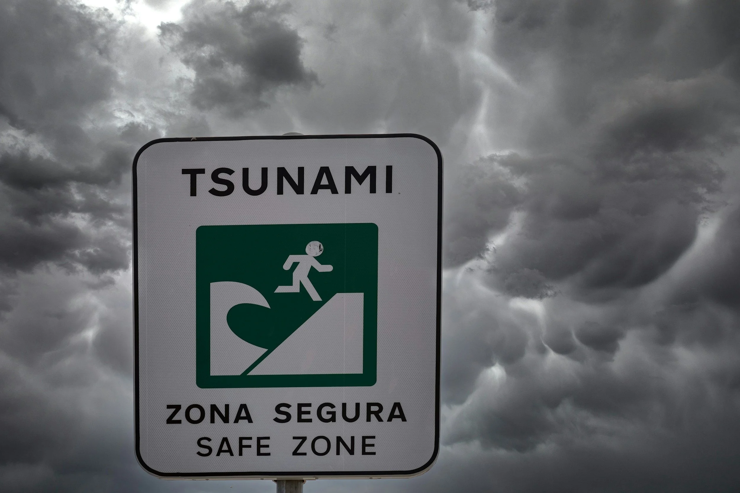 A warning sign indicating a tsunami safe zone with a person running from a large wave against a cloudy, stormy sky.