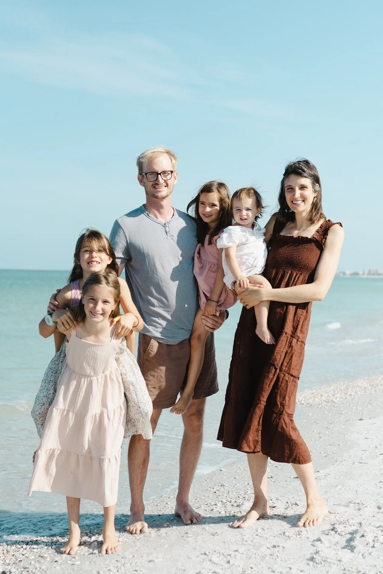 A family of six with three children, two women, and one man, standing on a beach with the ocean and sky in the background, smiling and enjoying a sunny day.