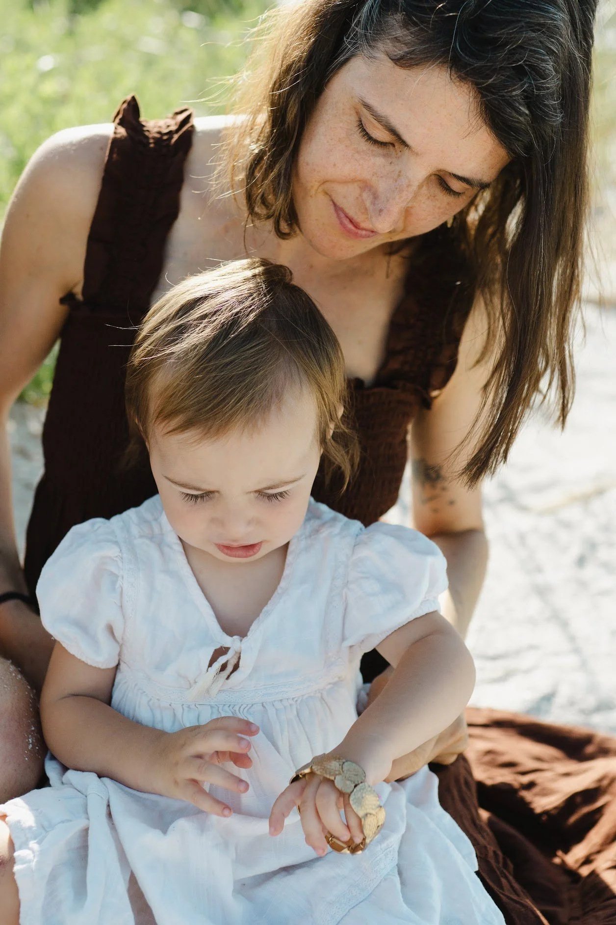 A woman with long brown hair and a sleeveless brown top is leaning over a young girl with light skin and short brown hair, who is wearing a white dress, sitting outdoors near water on a sunny day. The girl is looking closely at a small snake on her arm.