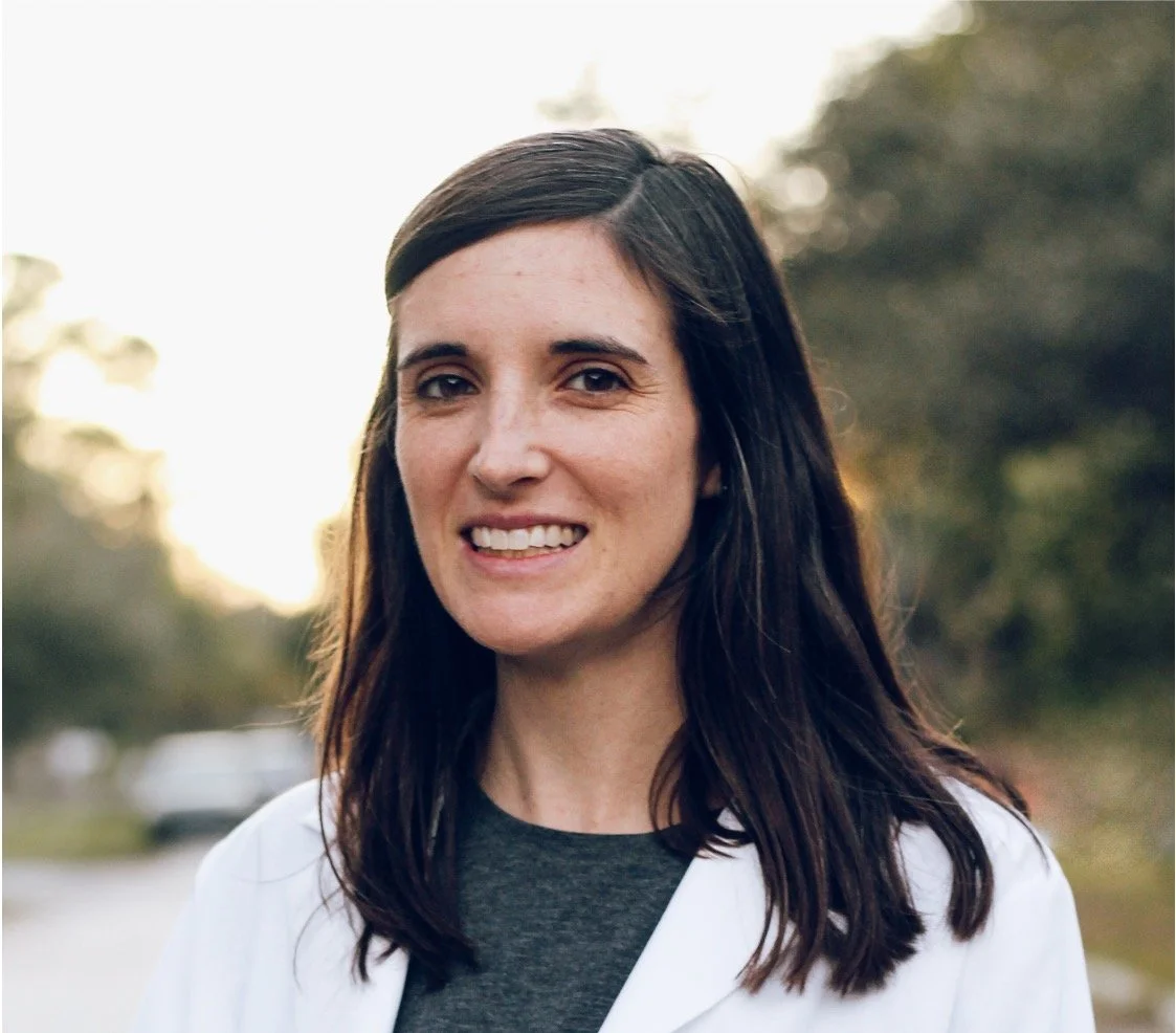 A woman with long dark hair smiling outdoors with trees and cars in the background.