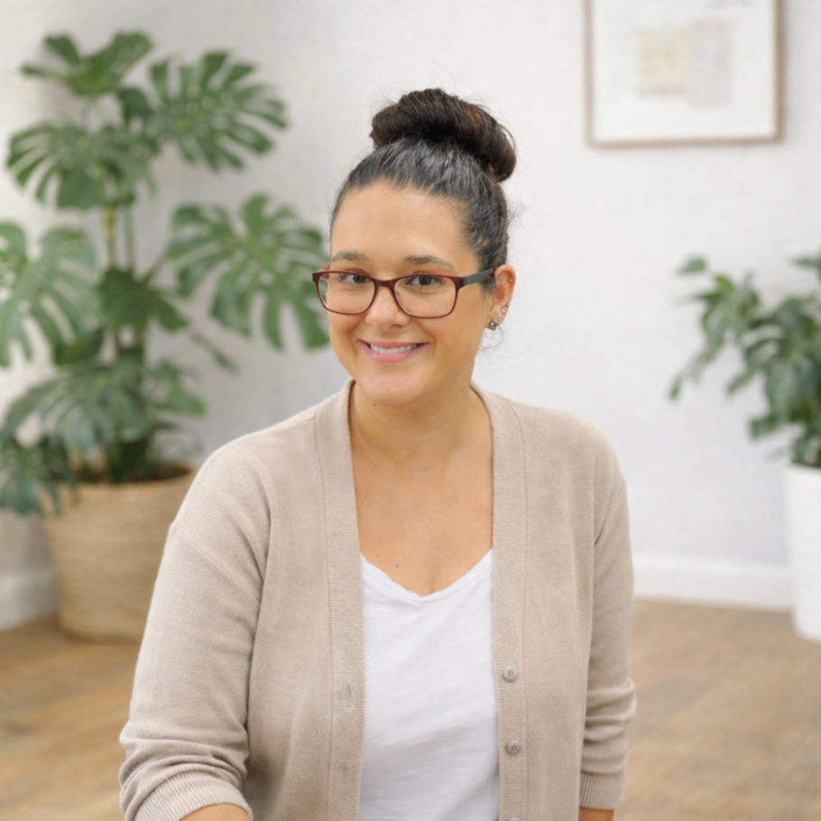 Smiling woman with dark hair in a bun, wearing glasses, a beige cardigan over a white top, sitting in a room with houseplants and framed pictures on the wall.
