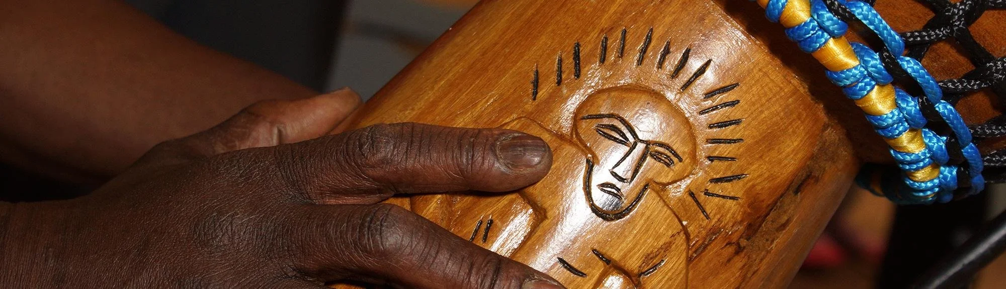 A person's hand holding a wooden object with a carved face of a man with sunglasses, surrounded by radiating lines, and wearing colorful braided bracelets.