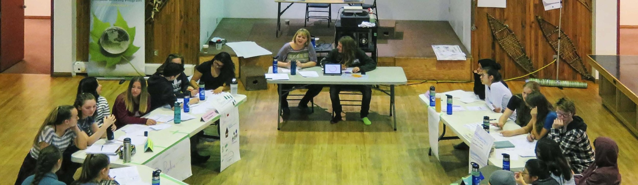 A group of young women seated at tables in a large room, possibly participating in a discussion or workshop. The room has wooden floors and walls, with some decorations and paper materials visible.