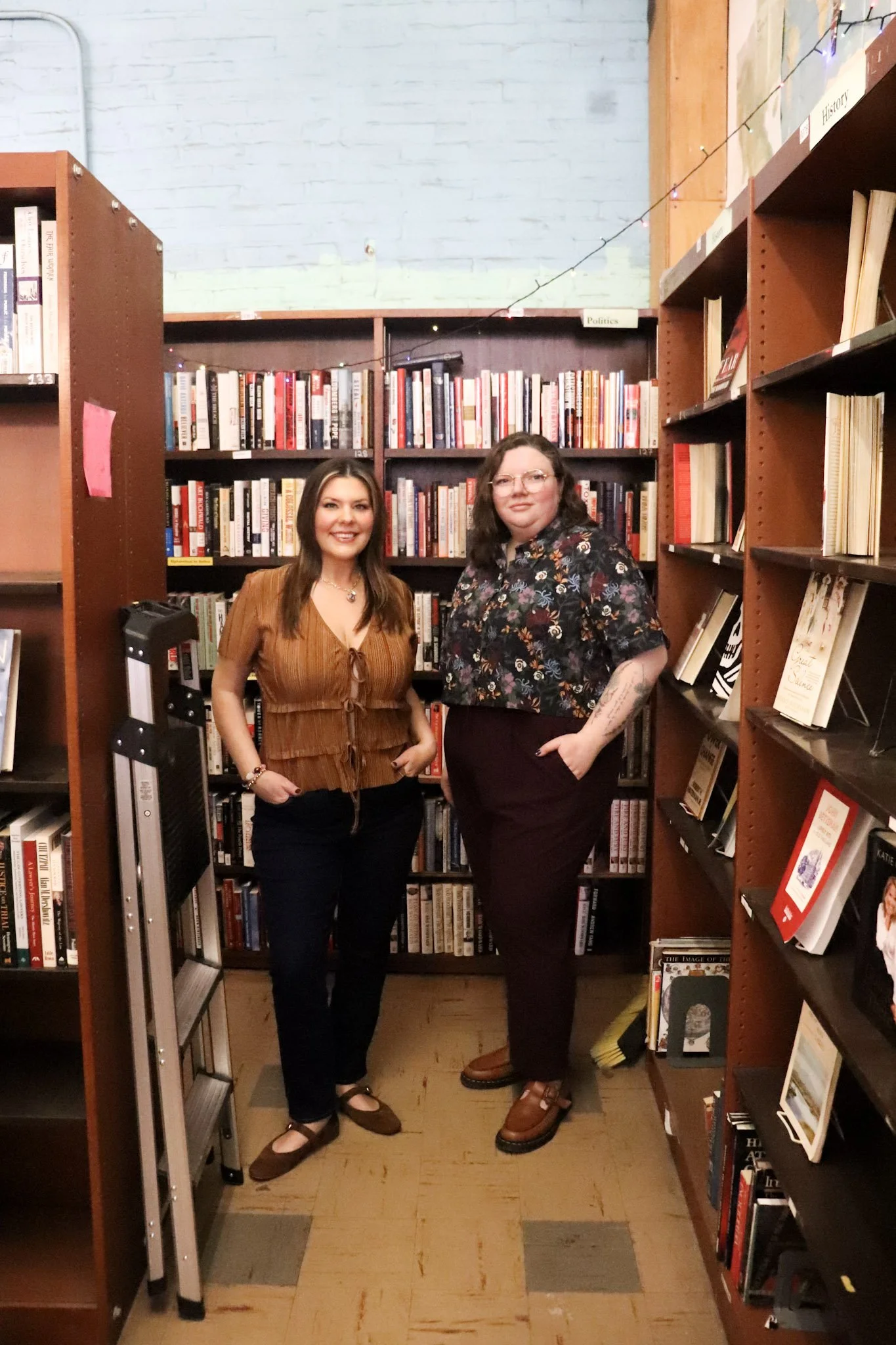 Two women standing in a bookstore aisle between bookshelves, smiling at the camera.