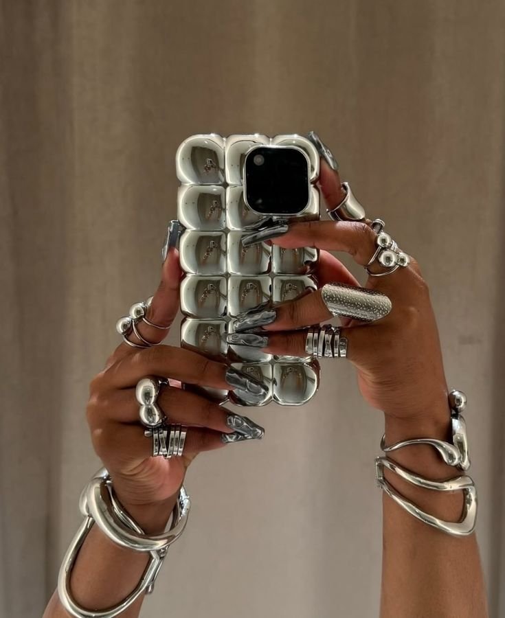 Person taking a mirror selfie with a silver, metallic, multi-fingered phone case and wearing multiple silver rings and bracelets.