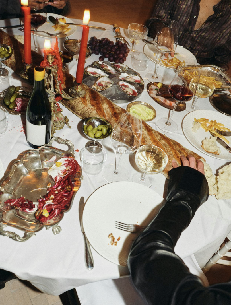 A dinner table set with wine glasses, plates, food, grapes, bread, and a centerpiece with candles. A person in a black leather jacket is reaching for a piece of bread.