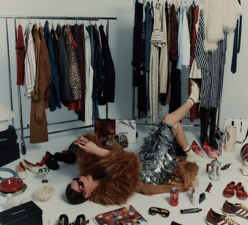 A woman lying on the floor surrounded by designer shopping bags, clothing, shoes, and accessories, with a clothing rack full of various garments behind her.