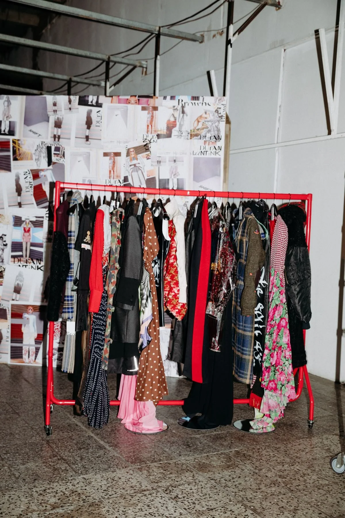 Clothing rack with various colorful fashion outfits and patterned fabrics, set against a wall covered with fashion magazine pages or posters.