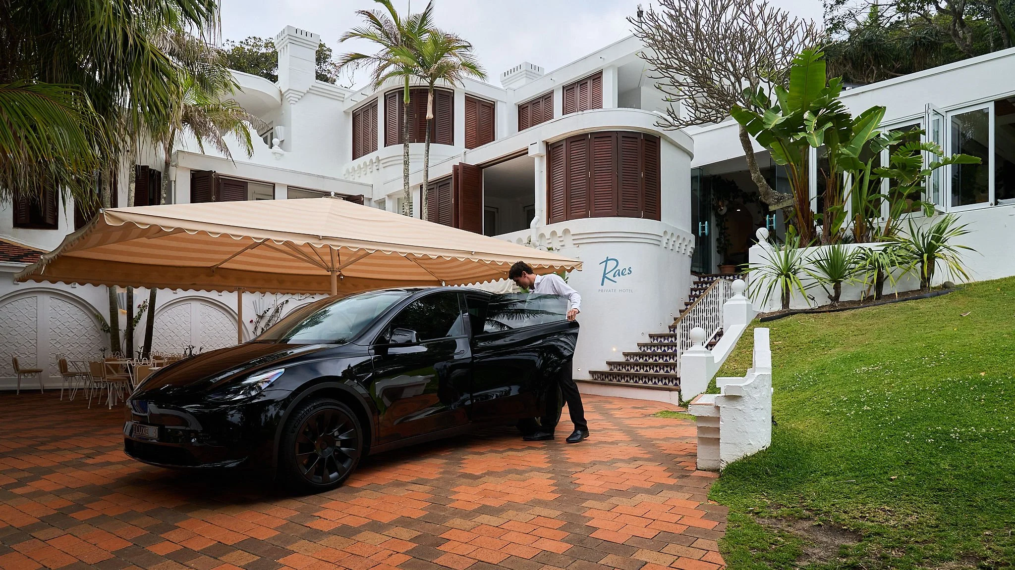 A man dressed in formal black pants and white shirt is loading a black car parked in front of a luxury white hotel named 'Raes Private Hotel'. The hotel has multiple levels with brown shutters on windows, a staircase leading up to the entrance, and surrounding greenery including palm trees. A beige umbrella covers part of an outdoor seating area on the brick-paved driveway.