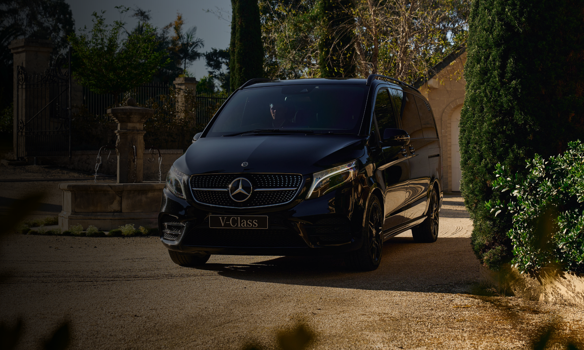 A black Mercedes-Benz V-Class van parked outdoors near greenery and a stone fountain in a courtyard.