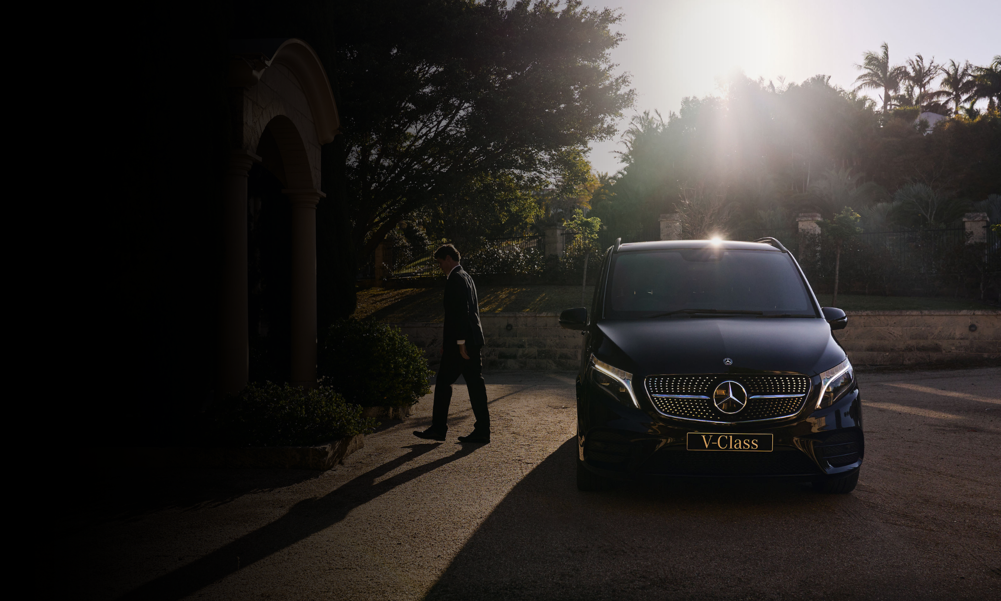 A man in a suit walking near a black Mercedes-Benz V-Class vehicle outdoors with sunlight filtering through trees in the background.