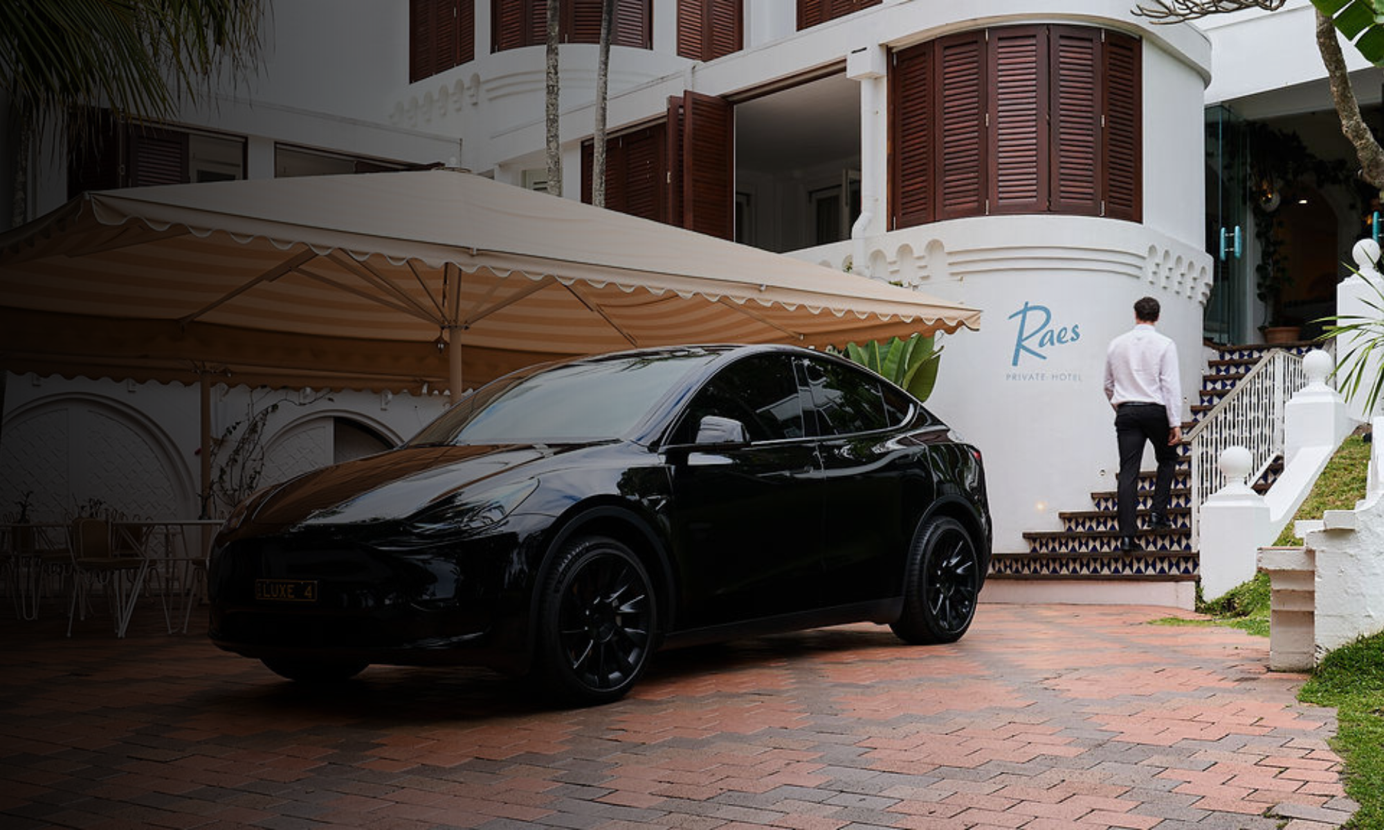 A man with dark hair wearing a white shirt and black pants walking up a staircase outside a white building with brown shutters. There is a black luxury car parked in front of the building under a beige striped canopy. The building has a sign that rea