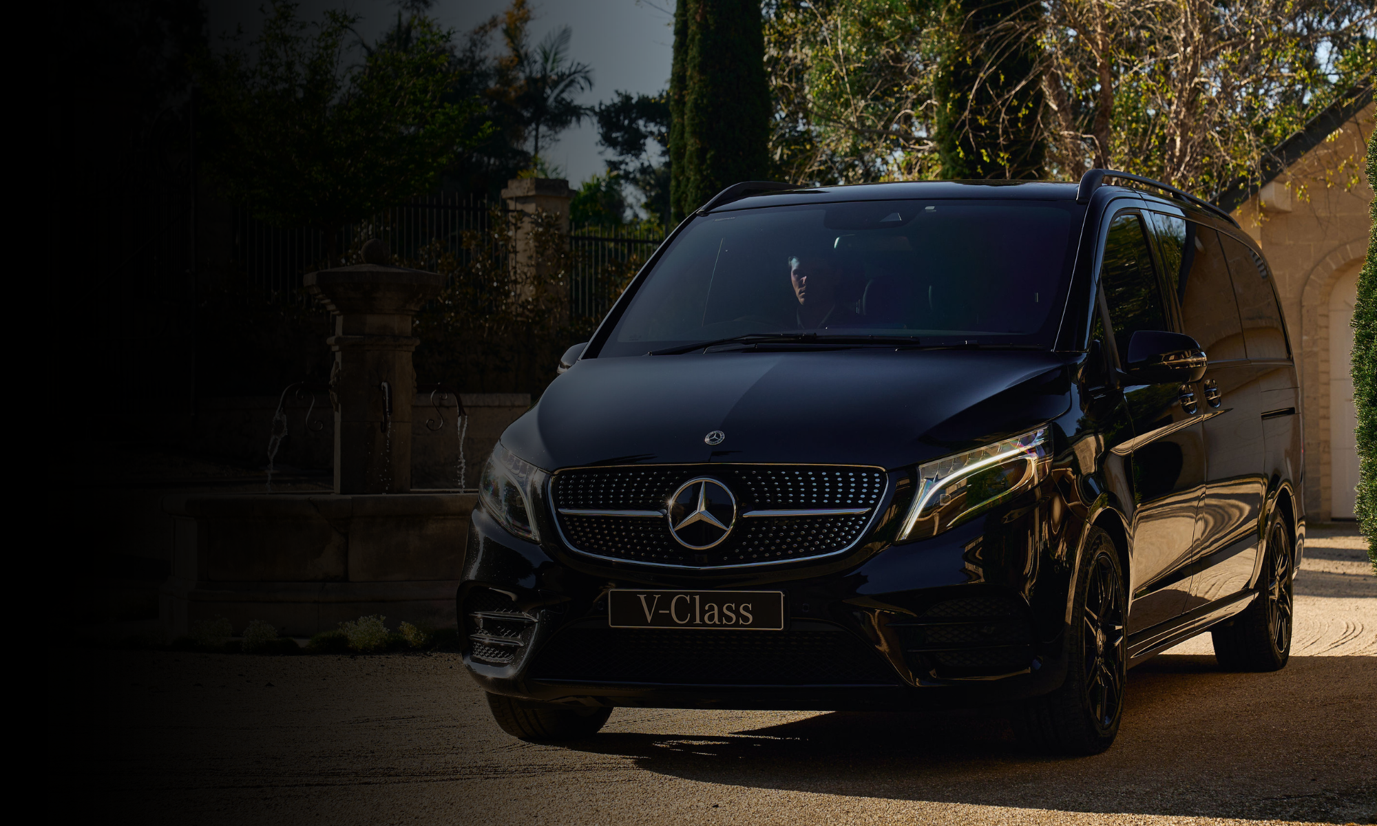 A black Mercedes-Benz V-Class minivan parked in a driveway with a fountain and trees in the background.