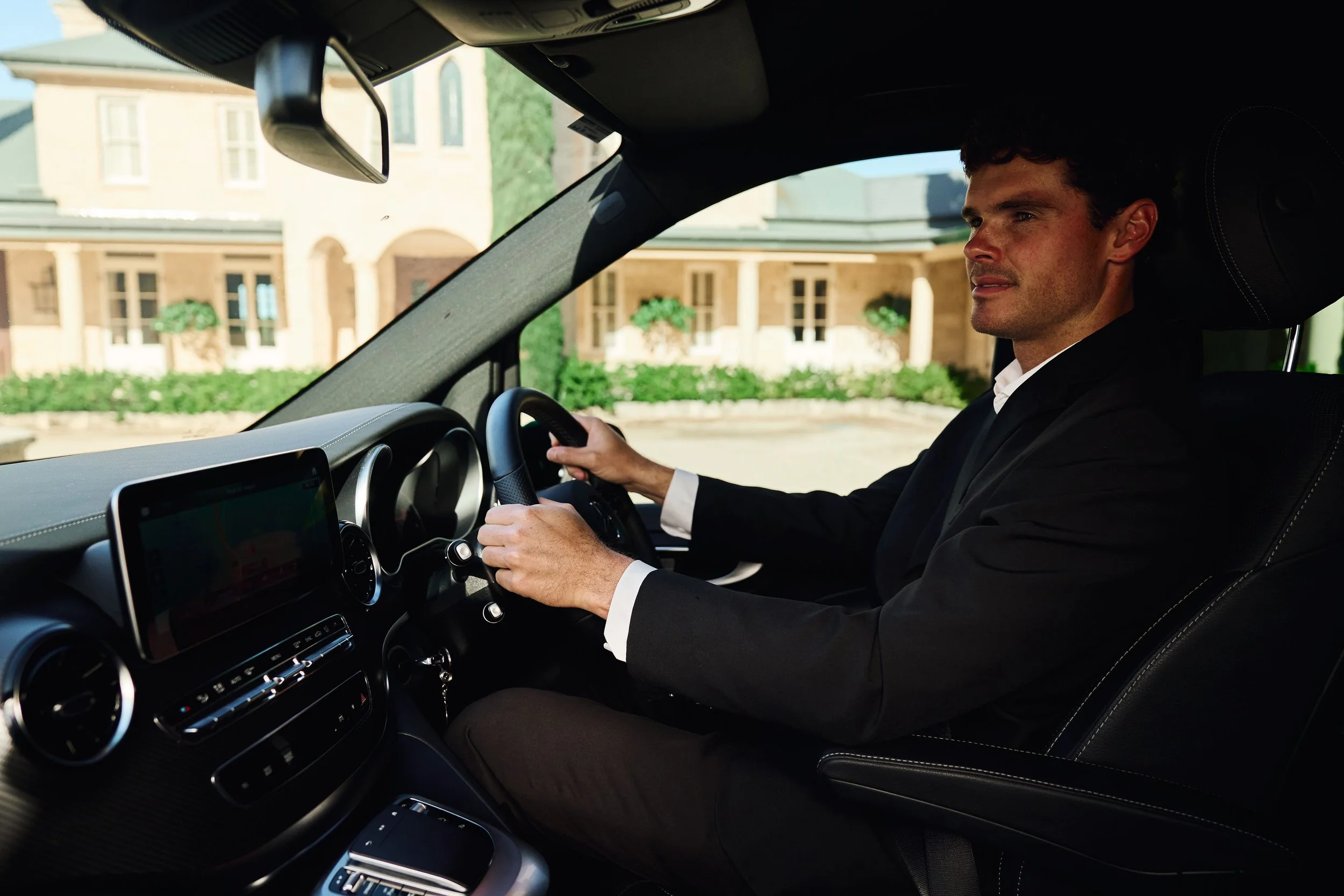 A man in a suit sitting in the driver's seat of a car, holding the steering wheel, with buildings visible through the windshield.