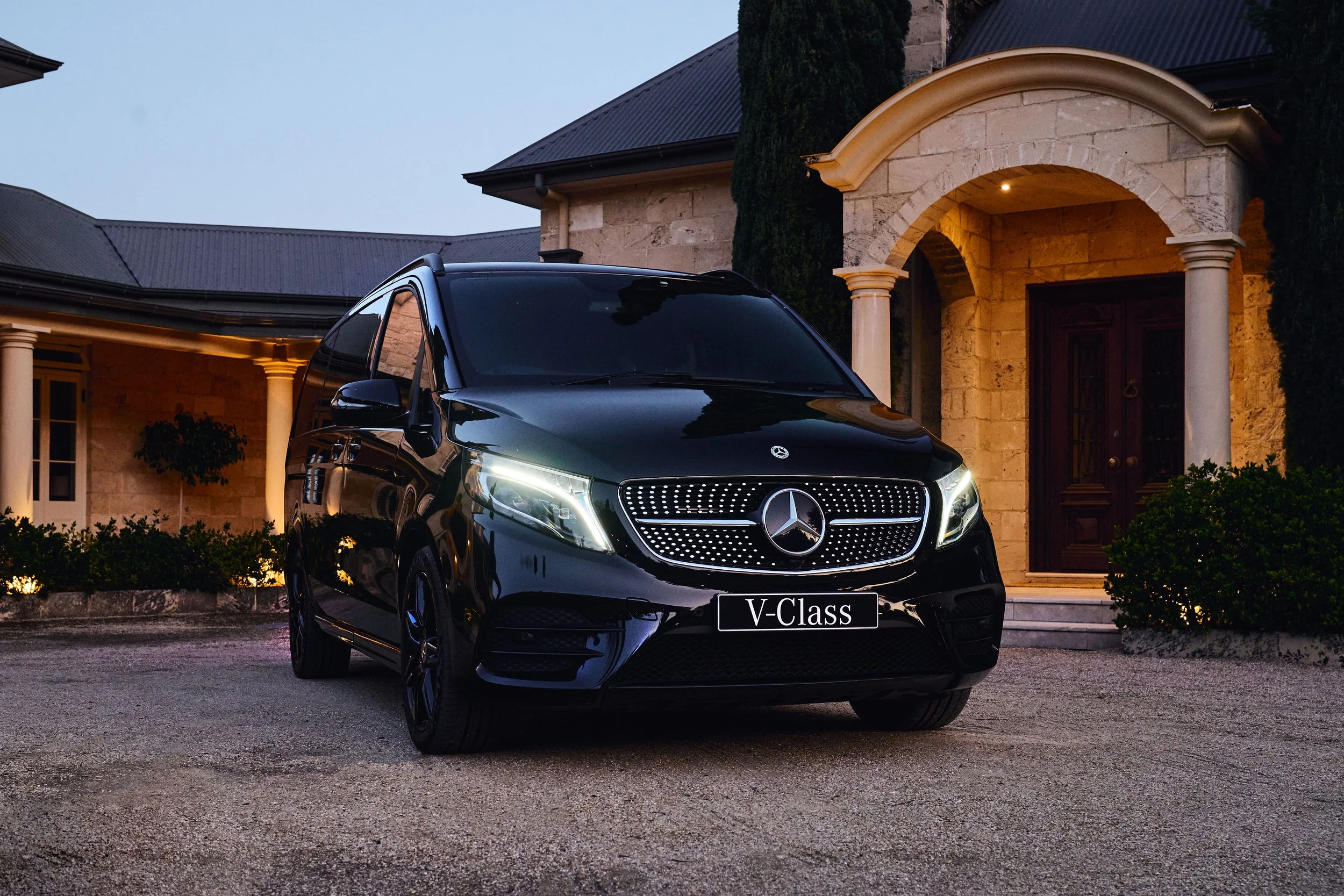 A black Mercedes-Benz V-Class van parked in front of a large stone house with illuminated entrance, bushes, and trees during evening.