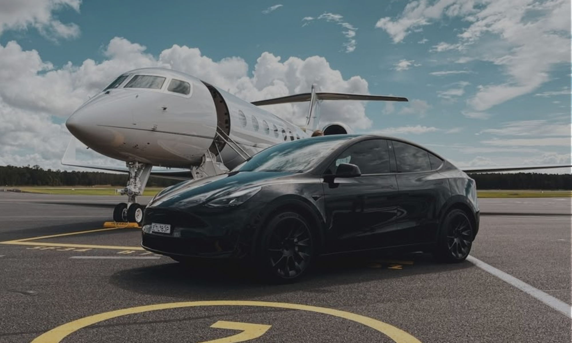 A black Tesla Model Y parked on an airport tarmac beneath a white private jet, with a partly cloudy blue sky in the background.