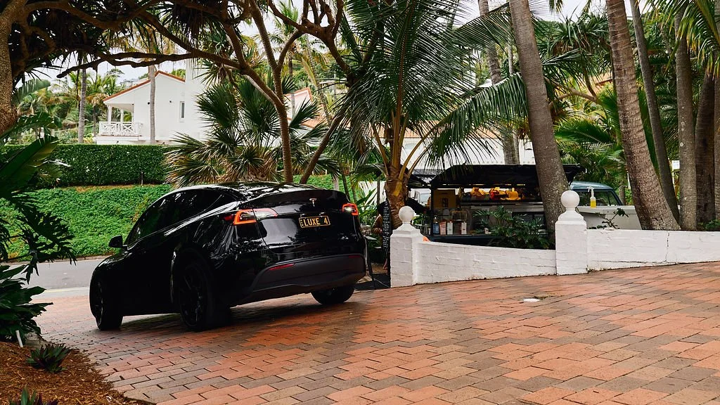 A sleek black Tesla parked on a brick driveway surrounded by tropical plants and trees, with a white wall and a small outdoor kitchen area in the background.