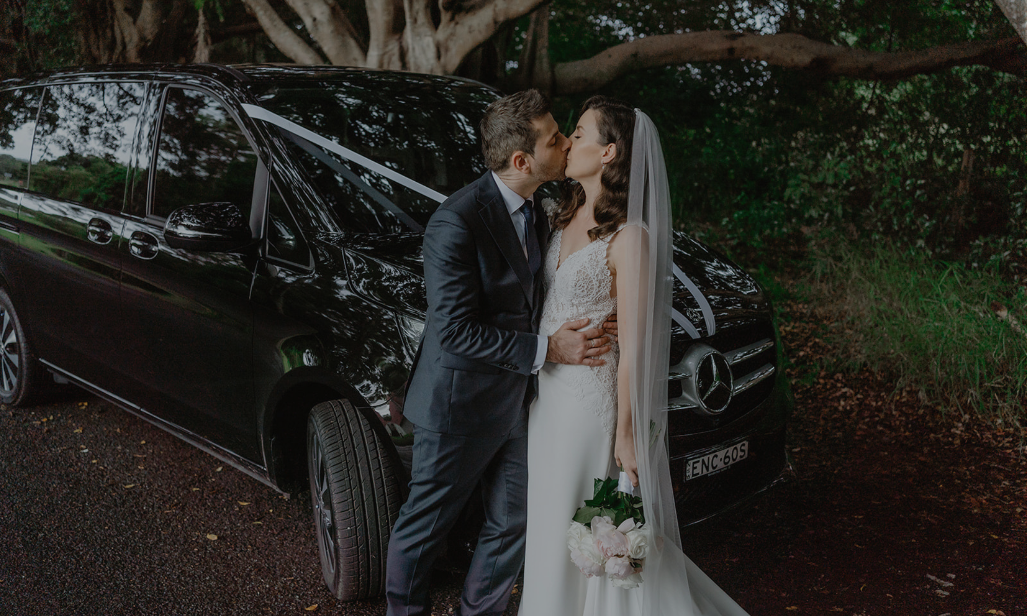 A bride and groom share a kiss in front of a black luxury car decorated with white ribbons, on a wooded outdoor setting during their wedding.