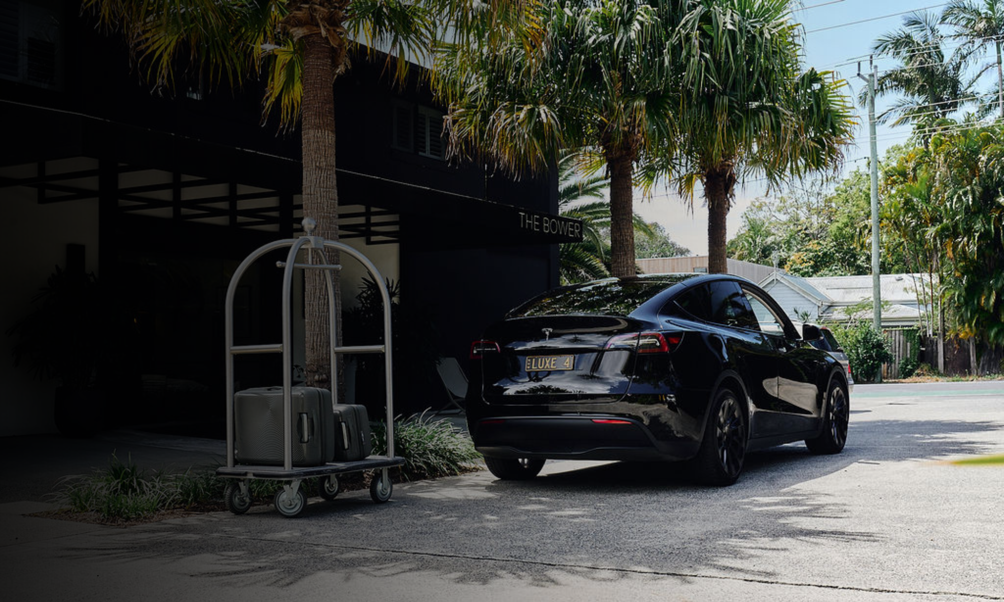 Black Tesla car parked outside a modern building with palm trees, luggage carts, and a sign reading "THE BOWER" in the background.