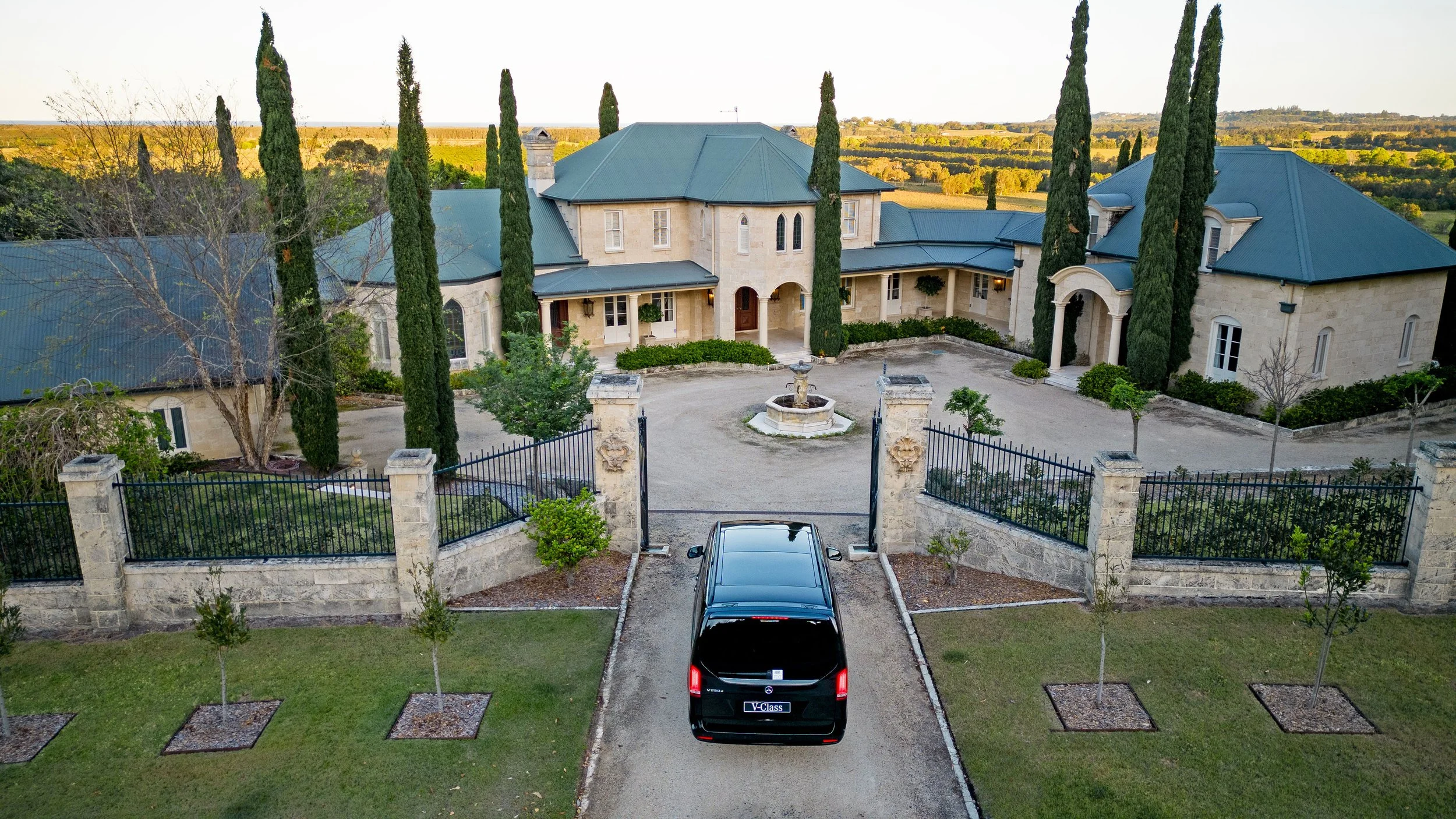A large mansion with a gated driveway, tall trees, a fountain, and a scenic countryside background.