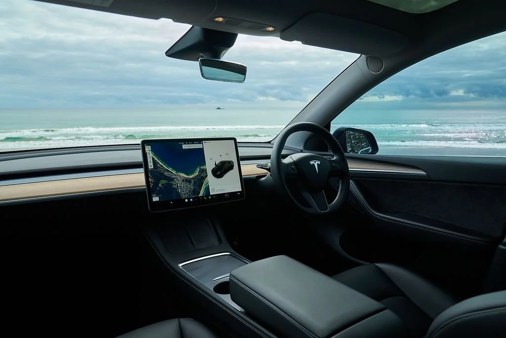 Inside a Tesla electric vehicle with a view of the ocean and cloudy sky through the front windshield.