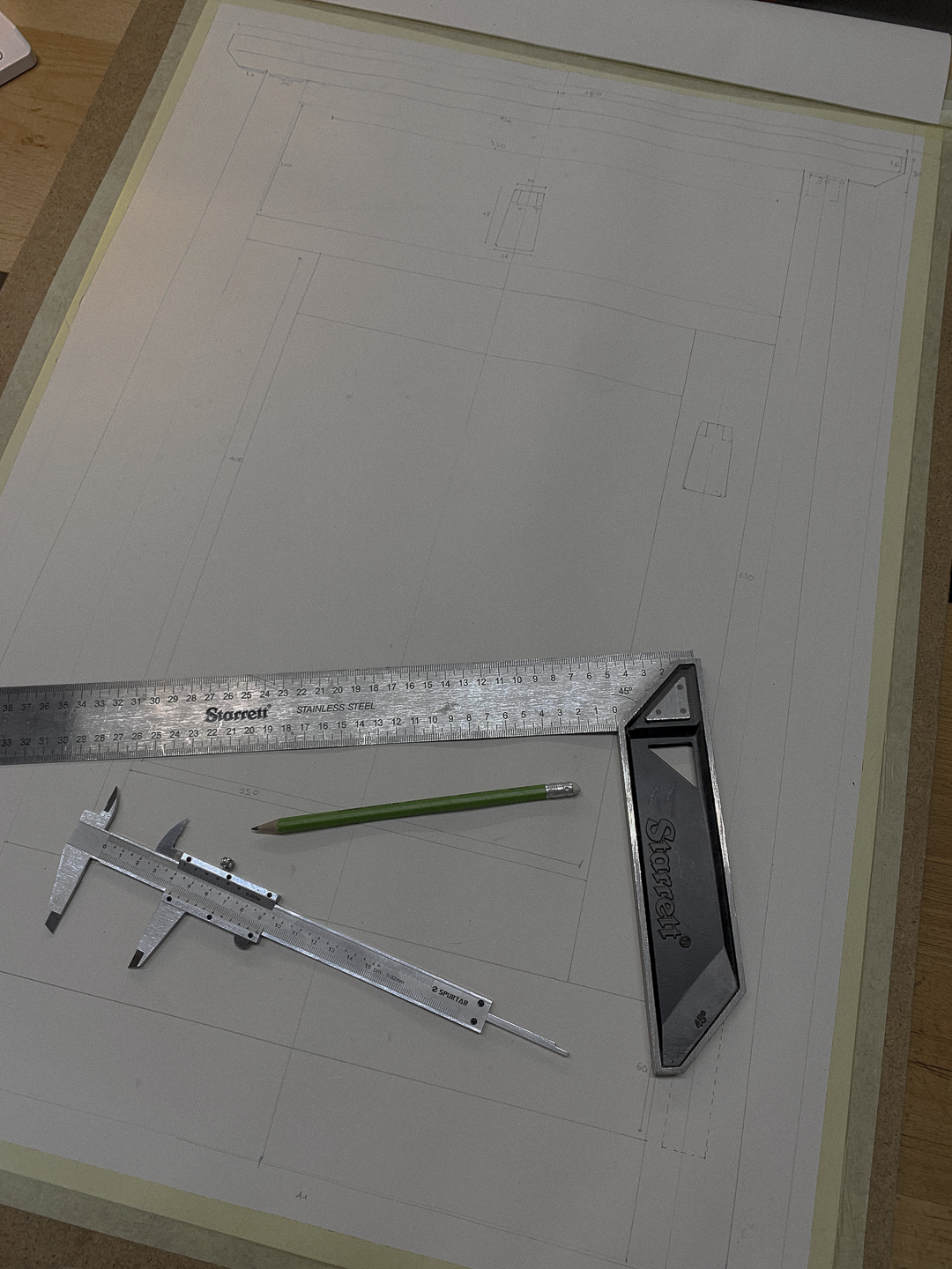 Blueprint drawing on a drafting table with a metal ruler, a green pencil, a sliding T-bevel, and a triangle ruler placed on it.