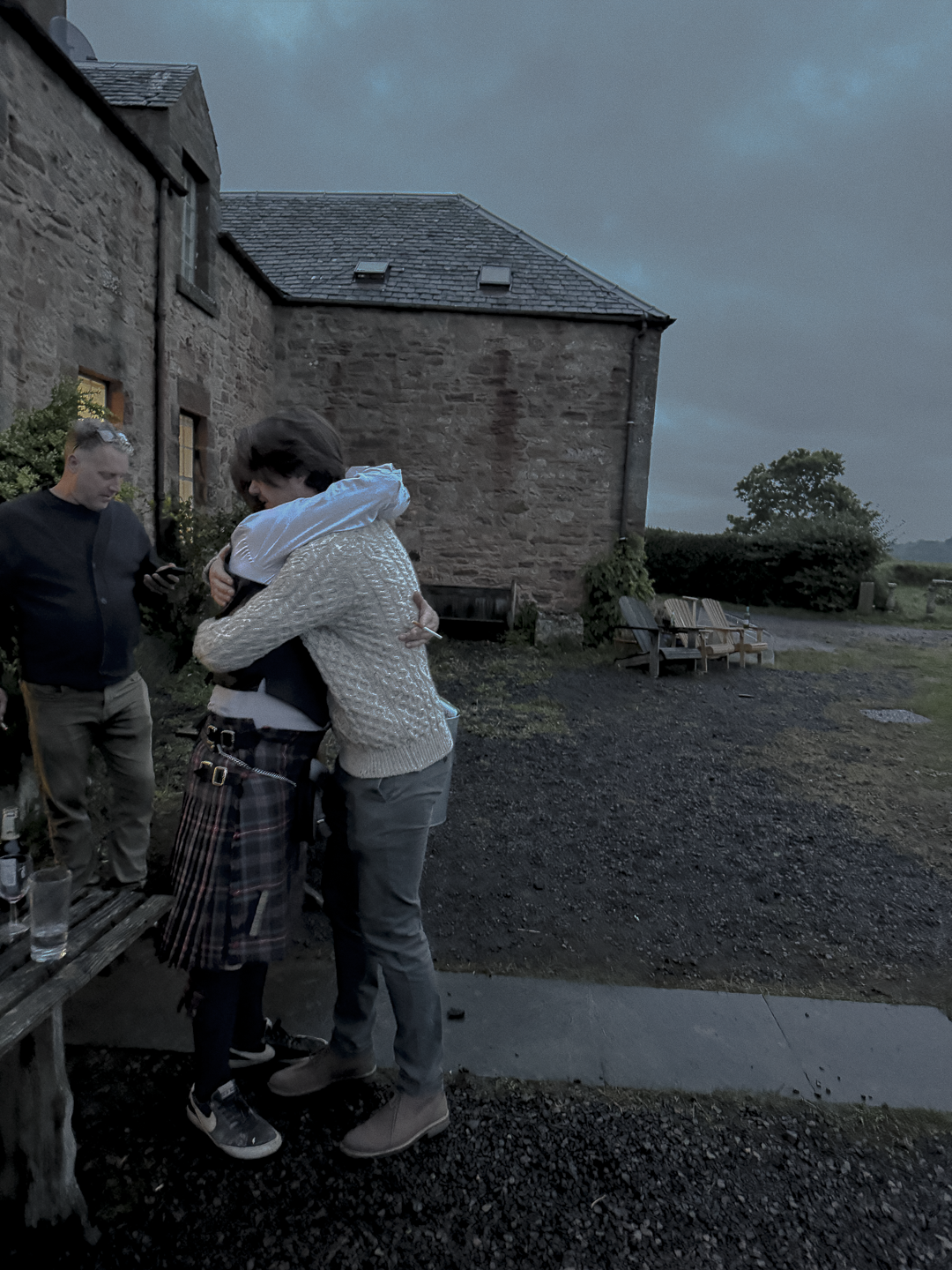 Two people embracing outdoors near a stone building during dusk, with another person standing nearby holding a phone; outdoor seating and a gravel path visible.