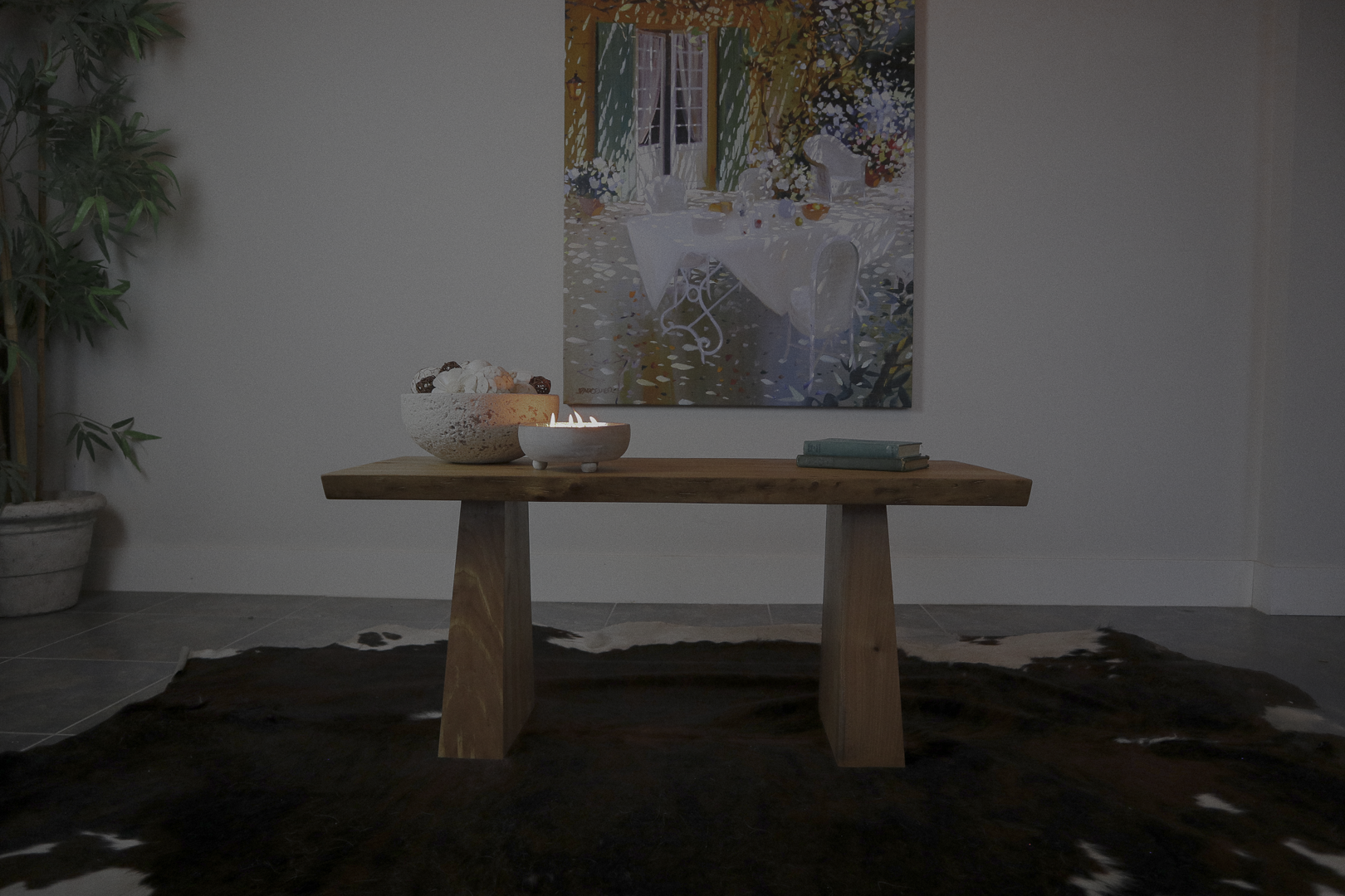A live edge wooden coffee table with a bowl of assorted decorations, a lit candle, and two books on it, placed in front of a colorful painting in a room with a dark cowhide rug and a potted plant.