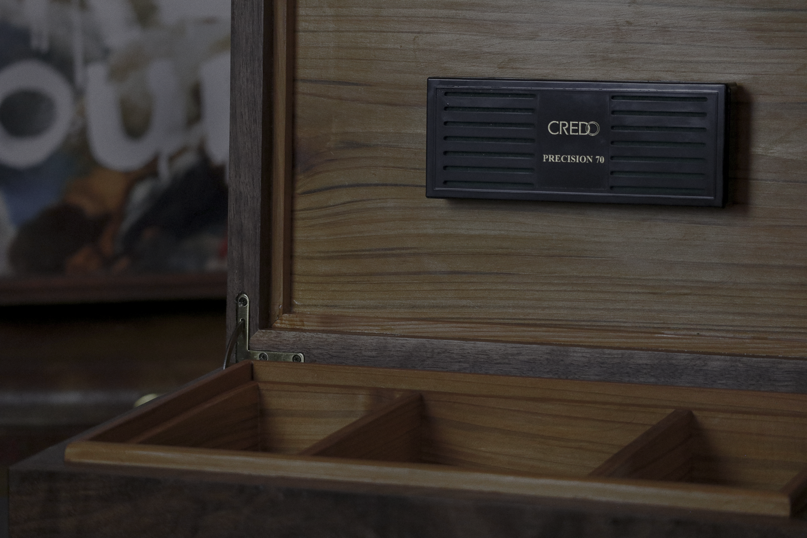 Close-up of an open wooden cigar humidor with a black Credo Precision 70 hygrometer mounted on the inside lid, showing elements of a blurred room in the background.