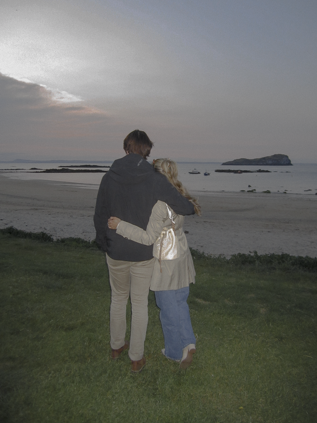 A couple with their arms around each other standing on a grassy area, looking at the ocean during sunset.