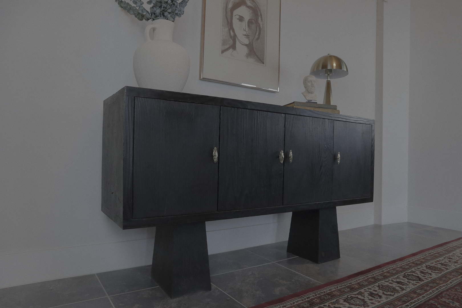 A black wooden sideboard with four doors, metallic handles, and angled legs, placed against a white wall. On top, there is a white vase with greenery, a framed sketch of a woman's face, a small bust sculpture, and a modern table lamp with a metallic shade. A patterned rug runs along the floor beside the sideboard.