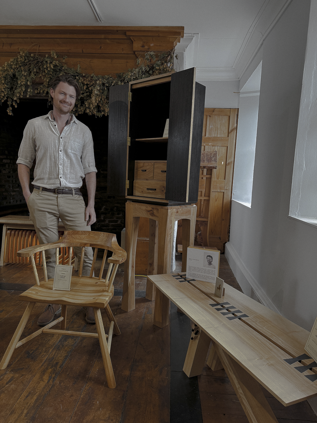 A man standing next to a display of handcrafted wooden furniture in a gallery setting the the Chippendale School of Furniture, with wooden chairs and a cabinet on display.