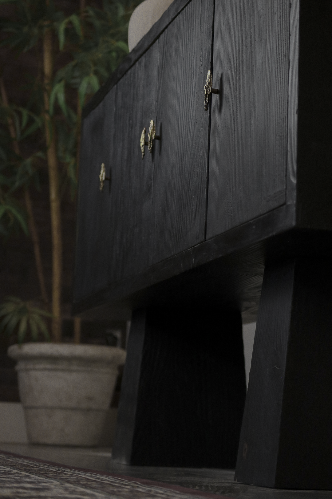 A black wooden side-board cabinet with small gold-colored geometric-shaped handles, standing on four wide legs in a room with a potted plant in the background.