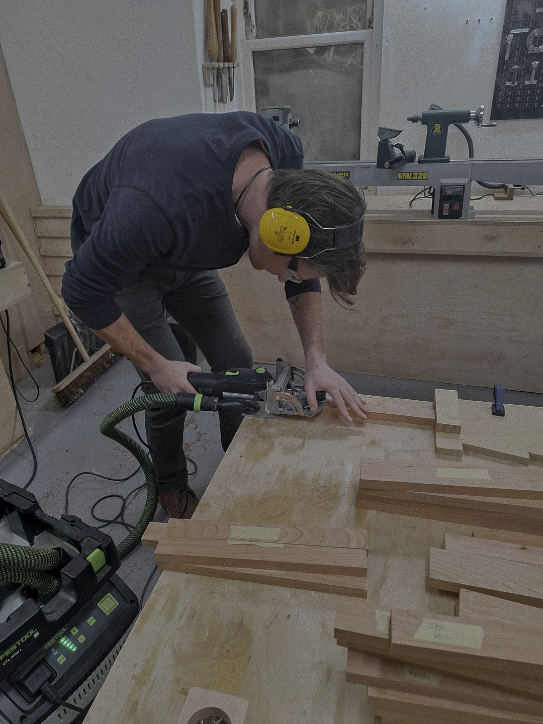 A man wearing yellow noise-canceling ear protection and glasses is cutting wood with a jigsaw in a workshop. Several pieces of wood are on the table, and tools are visible in the background.