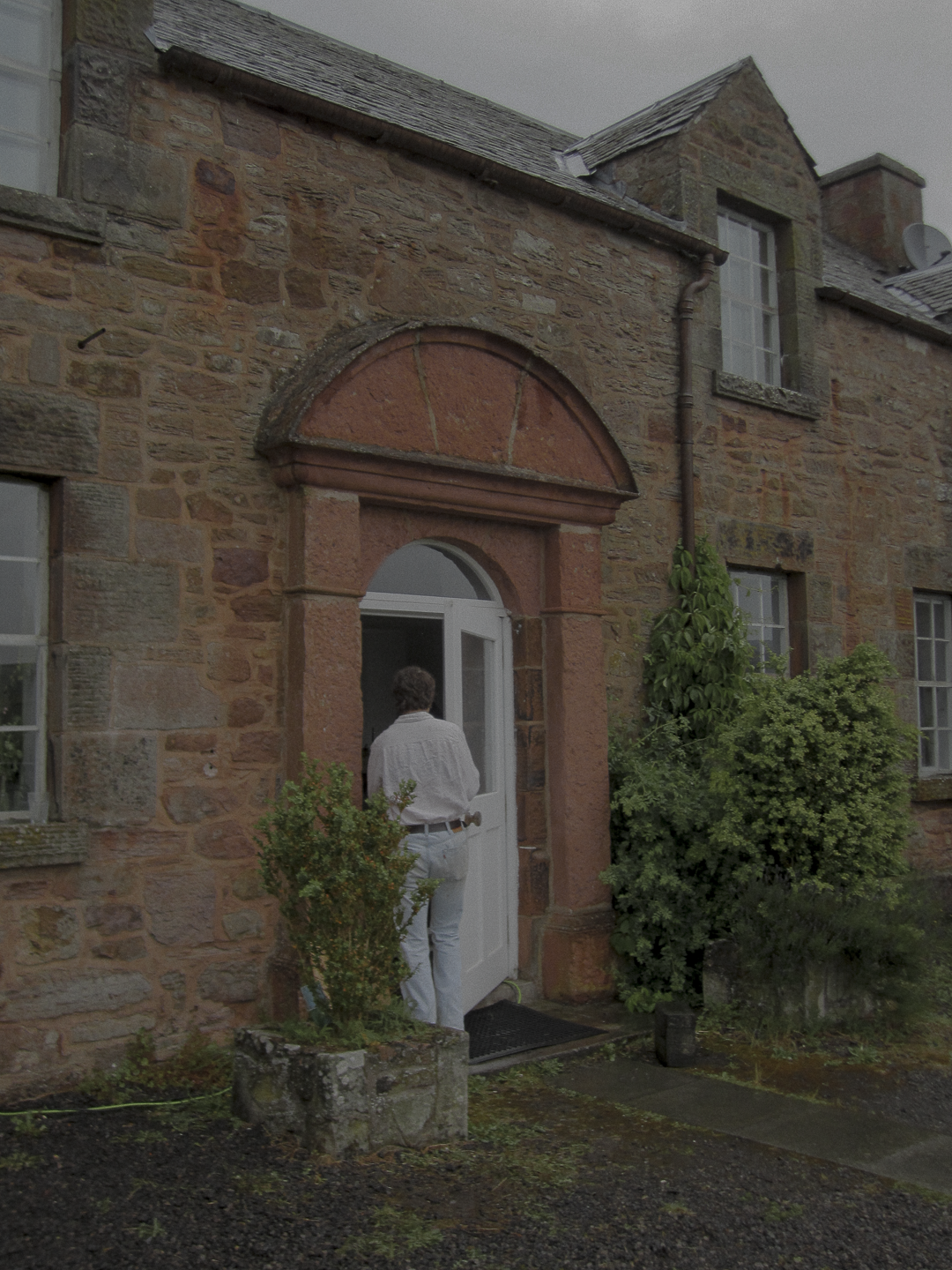 A person with curly hair wearing a white shirt and white pants opening a white door on a stone house with red and gray stones, surrounded by green bushes.