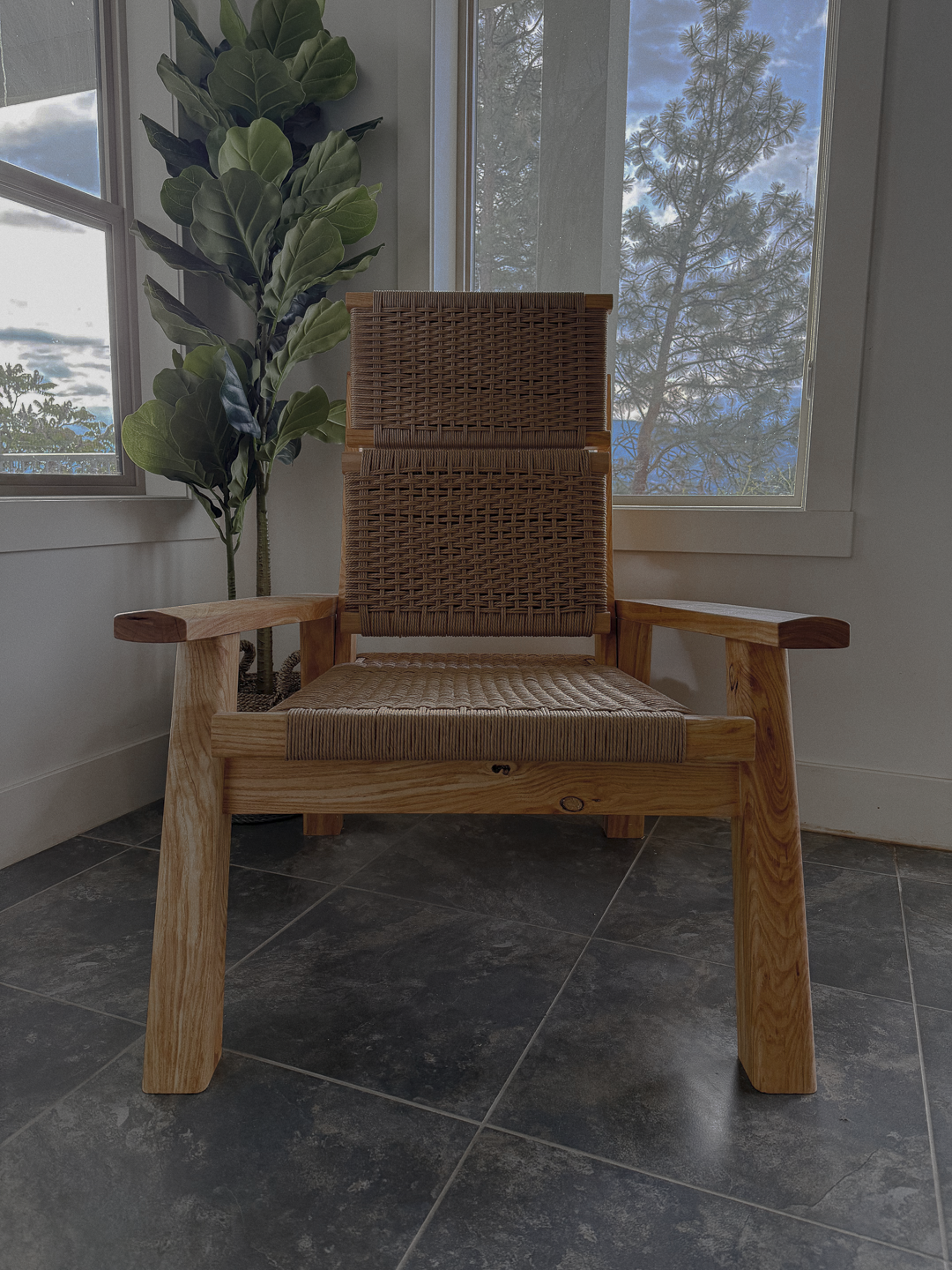 A woven wooden chair with a high back and padded seat, positioned on dark tiled flooring near large windows with a view of trees and a partly cloudy sky.