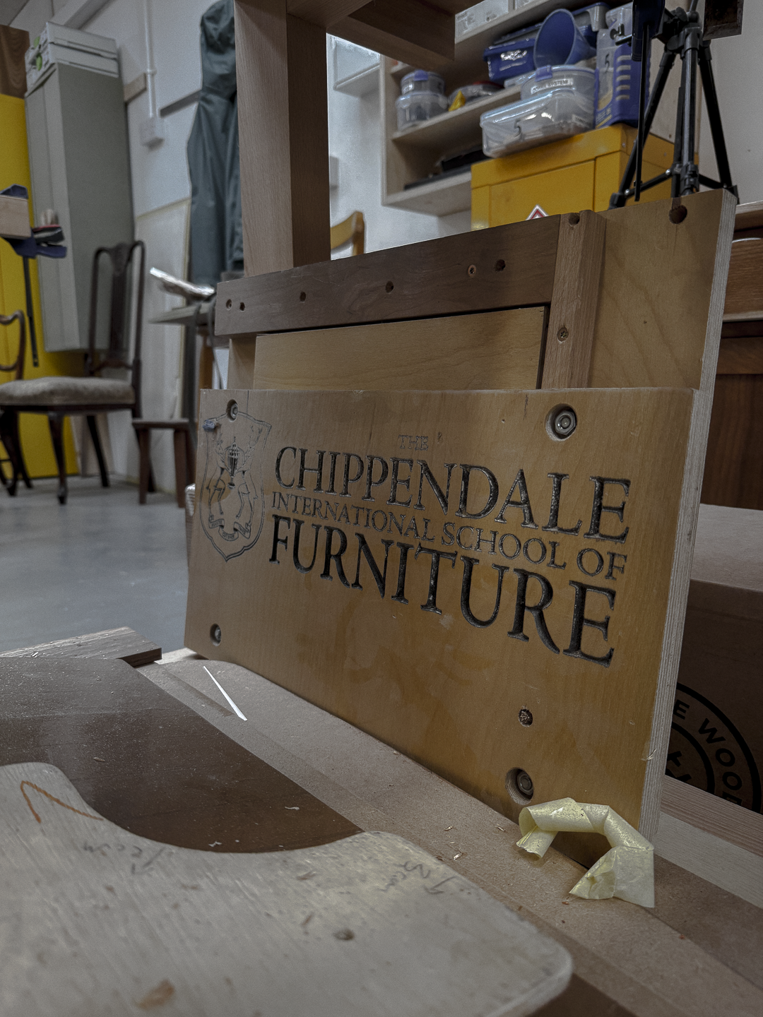 A wooden sign that reads 'The Chippendale International School of Furniture' on a workshop table with woodworking tools and furniture in the background.
