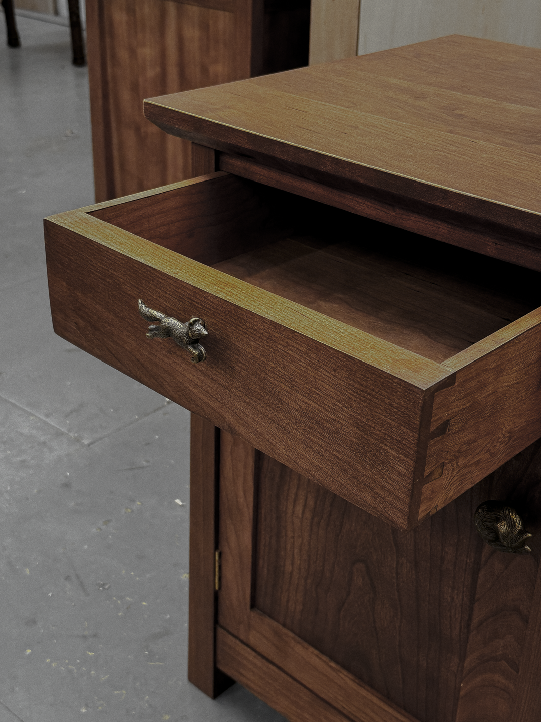 Close-up of a wooden bed-side dresser with an open drawer featuring a small fox-shaped handle, showing the detailed wood grain and dovetail joints, with more furniture in the background.